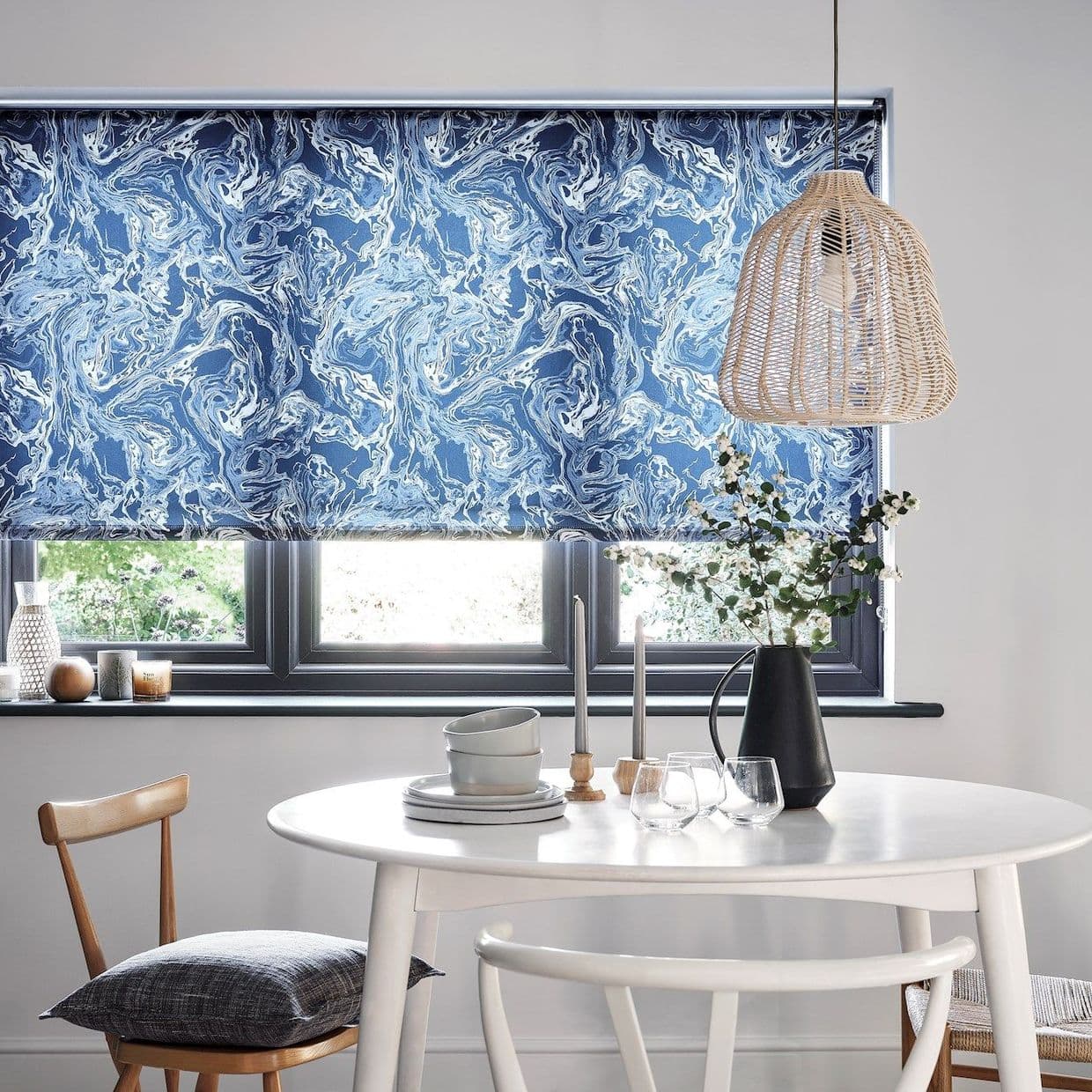 Rolled-down patterned roller blind, blue-and-white marbled, covering window and filtering daylight; bright dining nook with a white round table, wicker pendant lamp and a vase of branches.