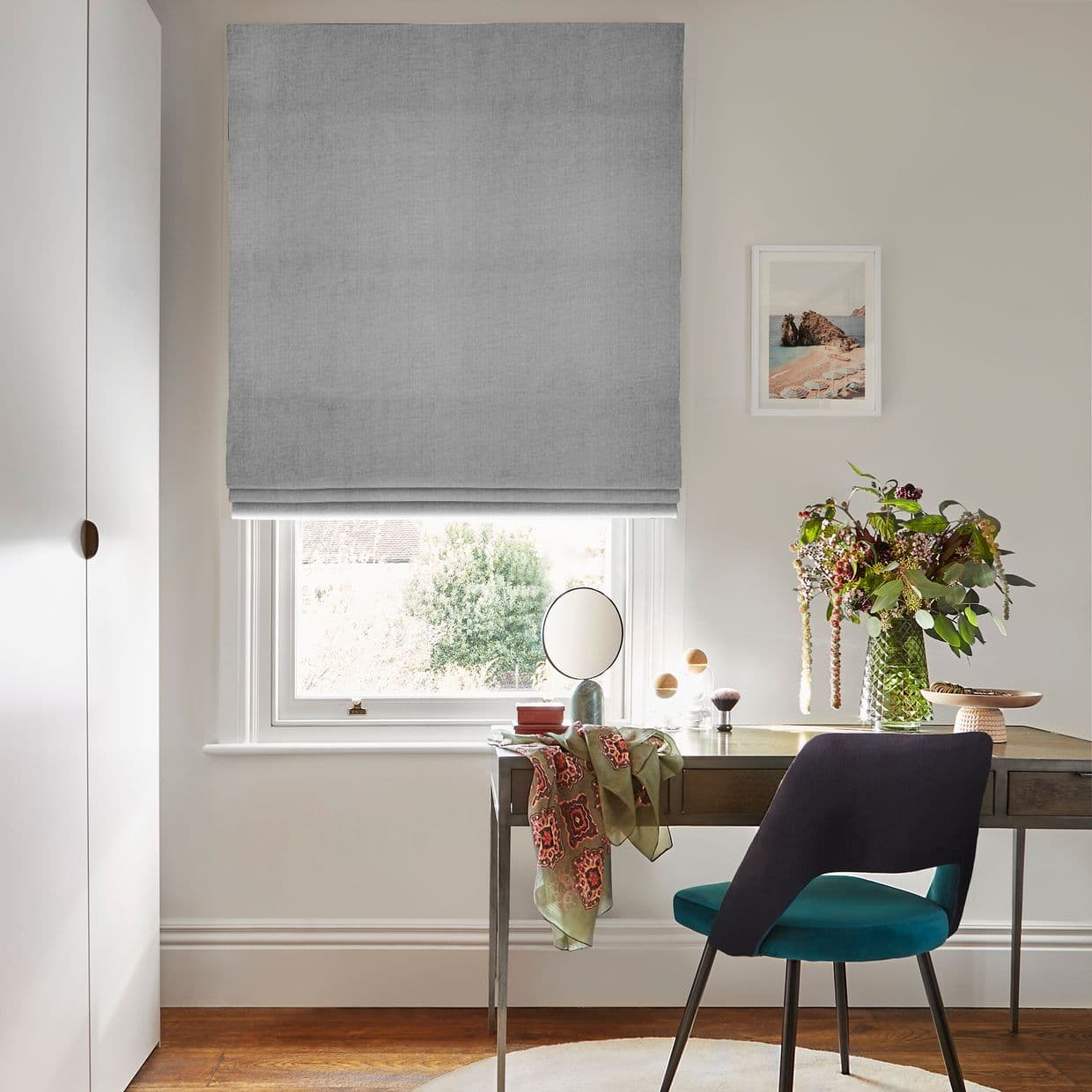 Grey Roman blind, lowered in soft folds, partially filtering daylight through the window; above a small desk in a bright home office with a teal chair and a vase of flowers.