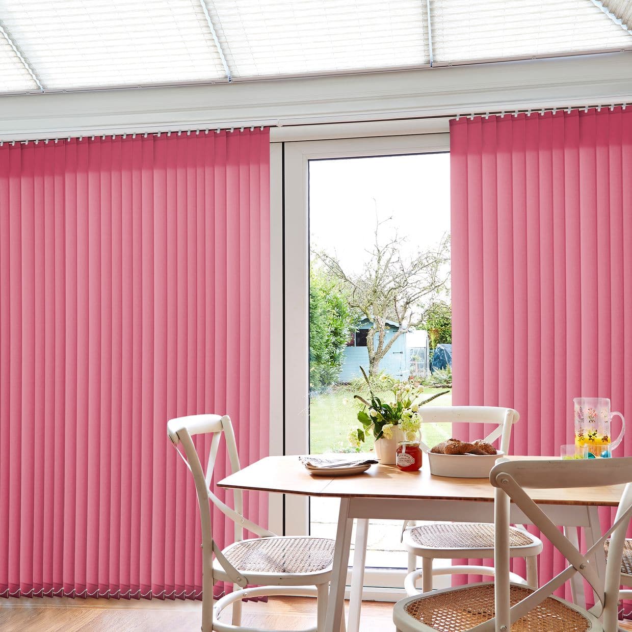 Pink vertical blinds, hung from a ceiling track, mostly closed with a central gap; filtering daylight into a bright dining area with wooden table, chairs and a garden view.
