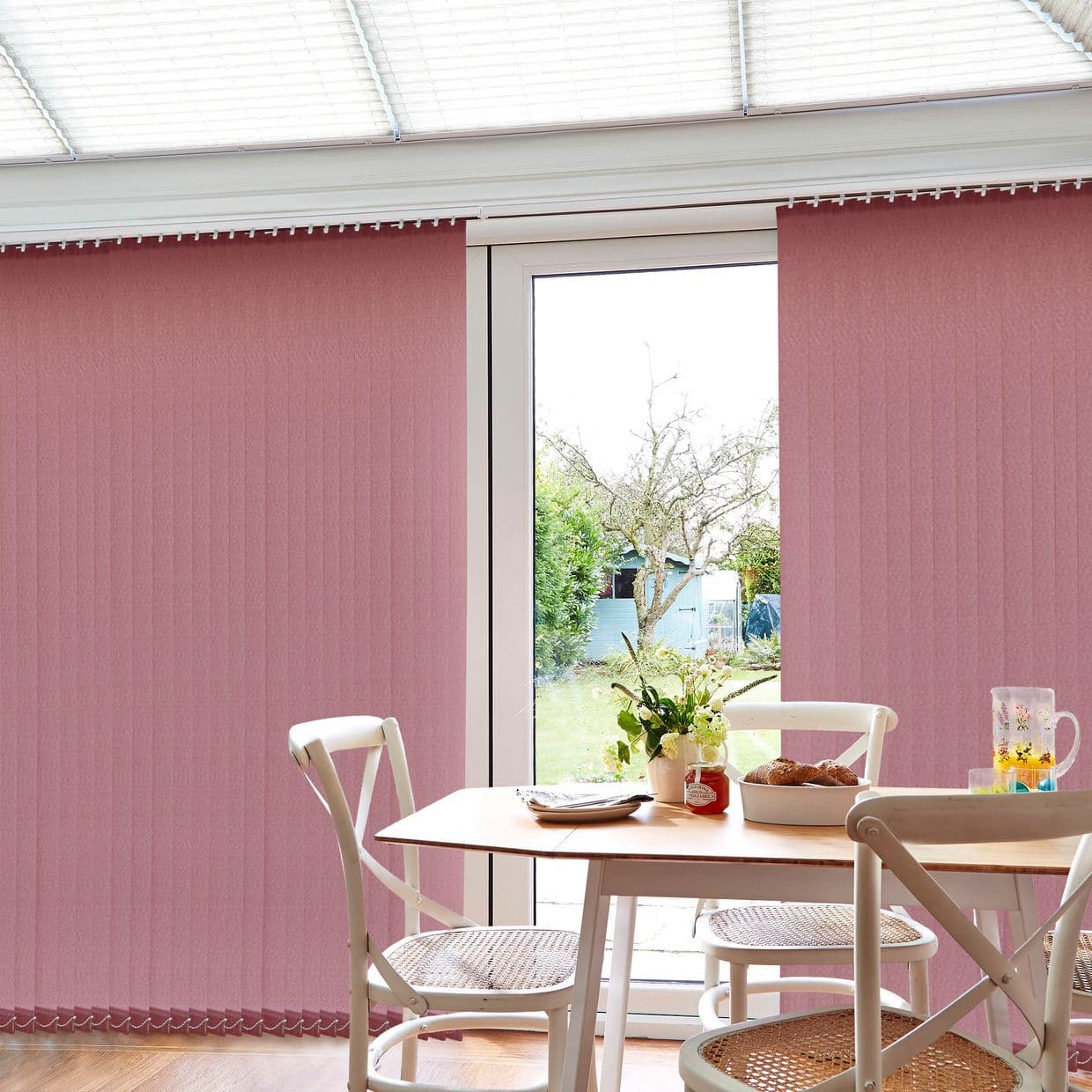 Pink vertical blinds cover a sliding glass door, panels mostly closed, filtering daylight with a central gap revealing a garden; small dining nook with wooden table, white chairs and a vase of flowers.