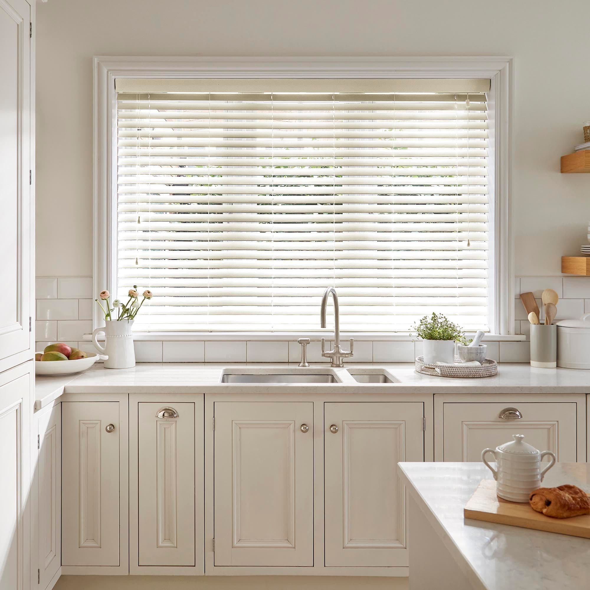 White horizontal Venetian blinds covering a large central window, partially tilted to filter bright daylight; framed by white trim above a cream kitchen sink and cabinetry with a light, minimalist countertop.