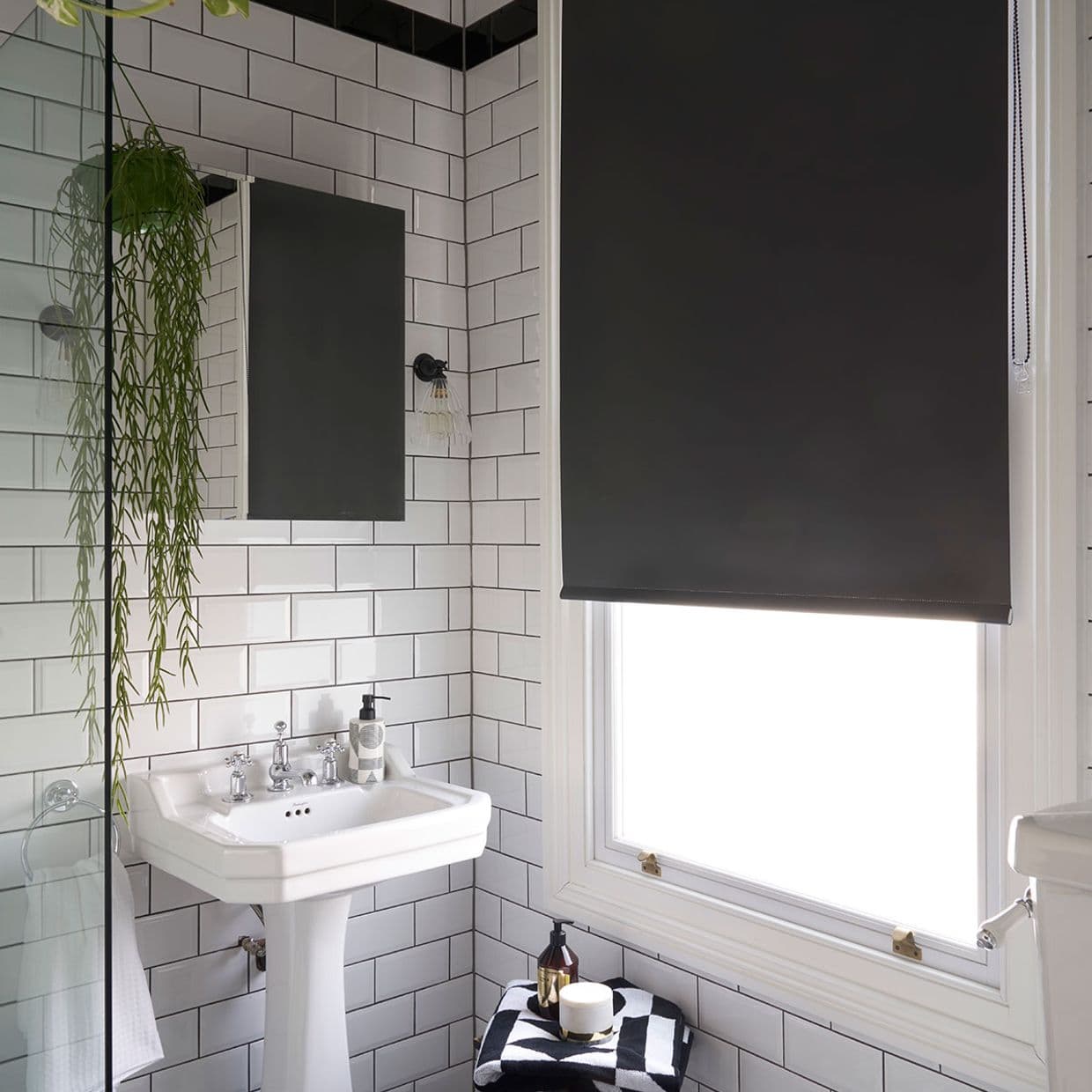Black roller blind, lowered with visible beaded pull, blocking light through a window; small white subway-tiled bathroom with pedestal sink, mirror, hanging plant, and toiletries on folded towel.
