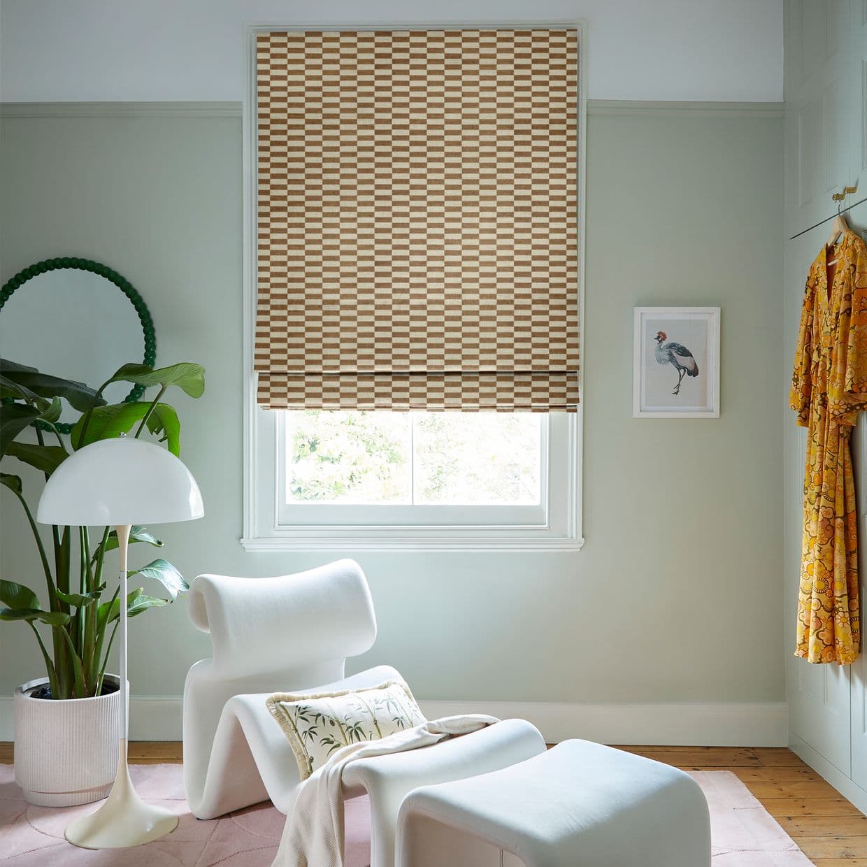 Beige-and-brown checkered Roman blind, lowered over the window and filtering daylight; set in a pale-green sitting nook with a white lounge chair, floor lamp, potted plant, and framed bird print.