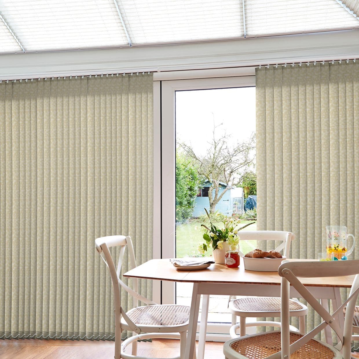 Beige vertical blinds covering a sliding glass patio door, panels mostly closed but parted at center; filtering daylight into a breakfast nook with table, chairs, vase, and garden view.