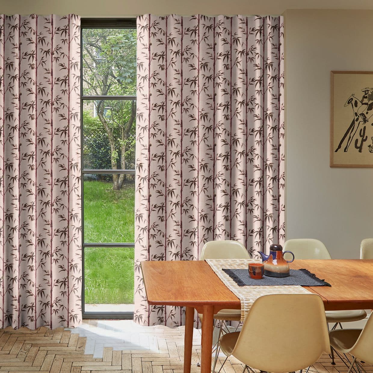 Floor-length pleated curtains, pink with brown bamboo print, partly drawn across a window; they filter daylight into a dining room with wooden table, beige chairs, parquet floor, grassy backyard.