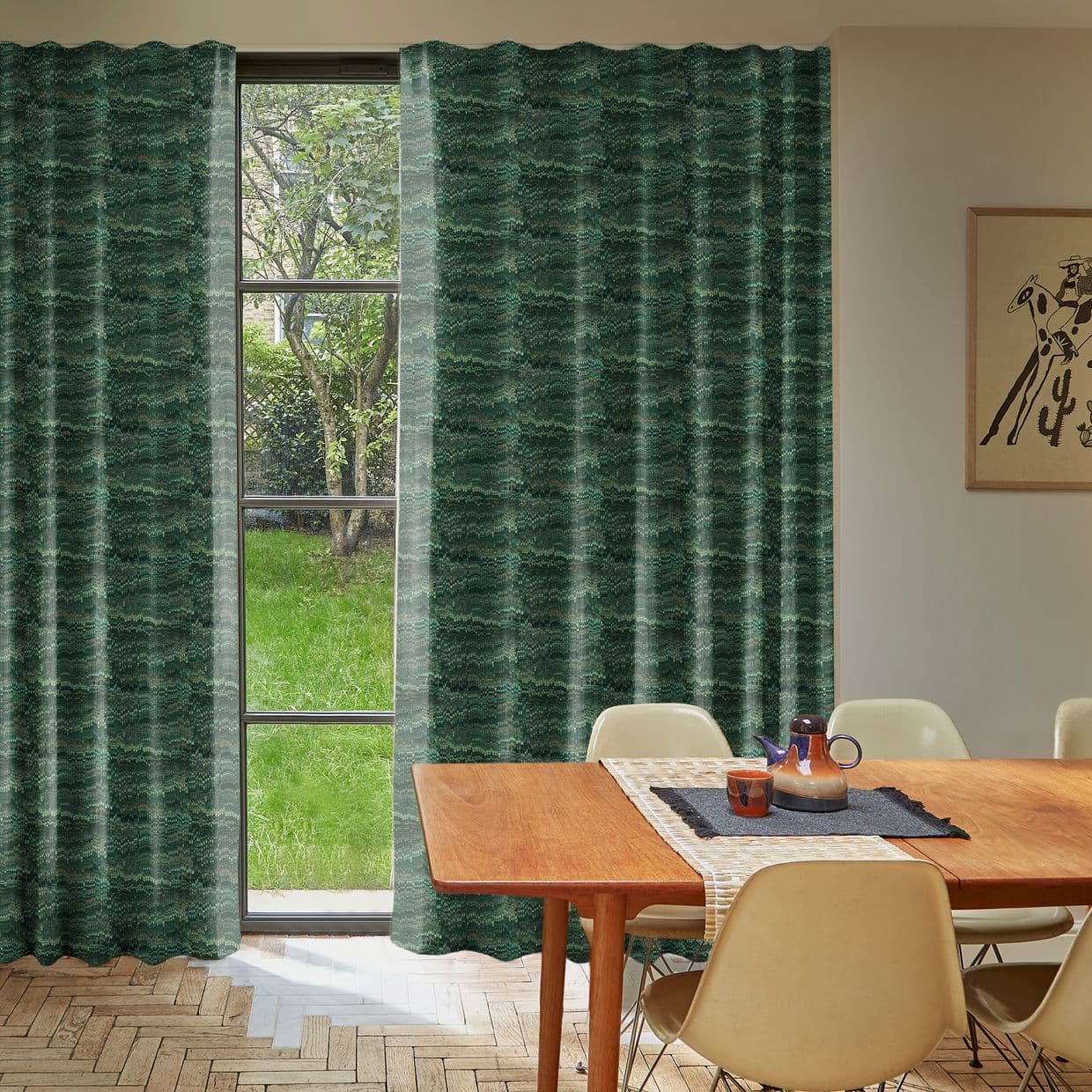 Green patterned, floor-length pleated curtains, mostly closed with a narrow central parting, filtering daylight and revealing a grassy backyard tree; mid-century dining room table, beige chairs and teapot nearby.