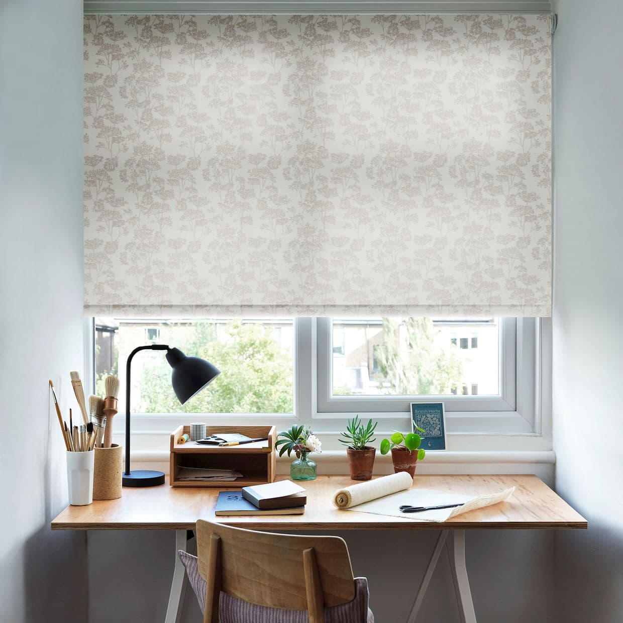 Floral-patterned roller blind, lowered and filtering soft daylight; wooden desk beneath holds a lamp, brushes, notebooks and potted succulents, with a tree-lined view outside.