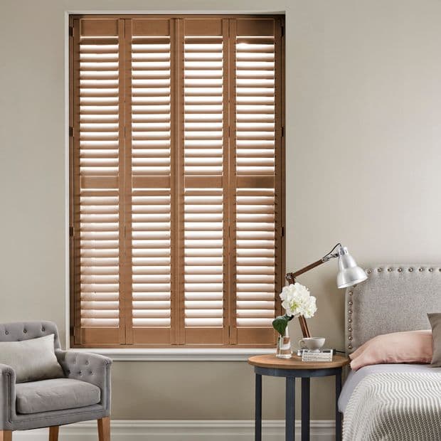 Wooden plantation shuttersâ€”four-panel, louveredâ€”cover a tall window, slats partially open and filtering light; bedroom corner with upholstered bed, bedside table, lamp, and gray armchair.