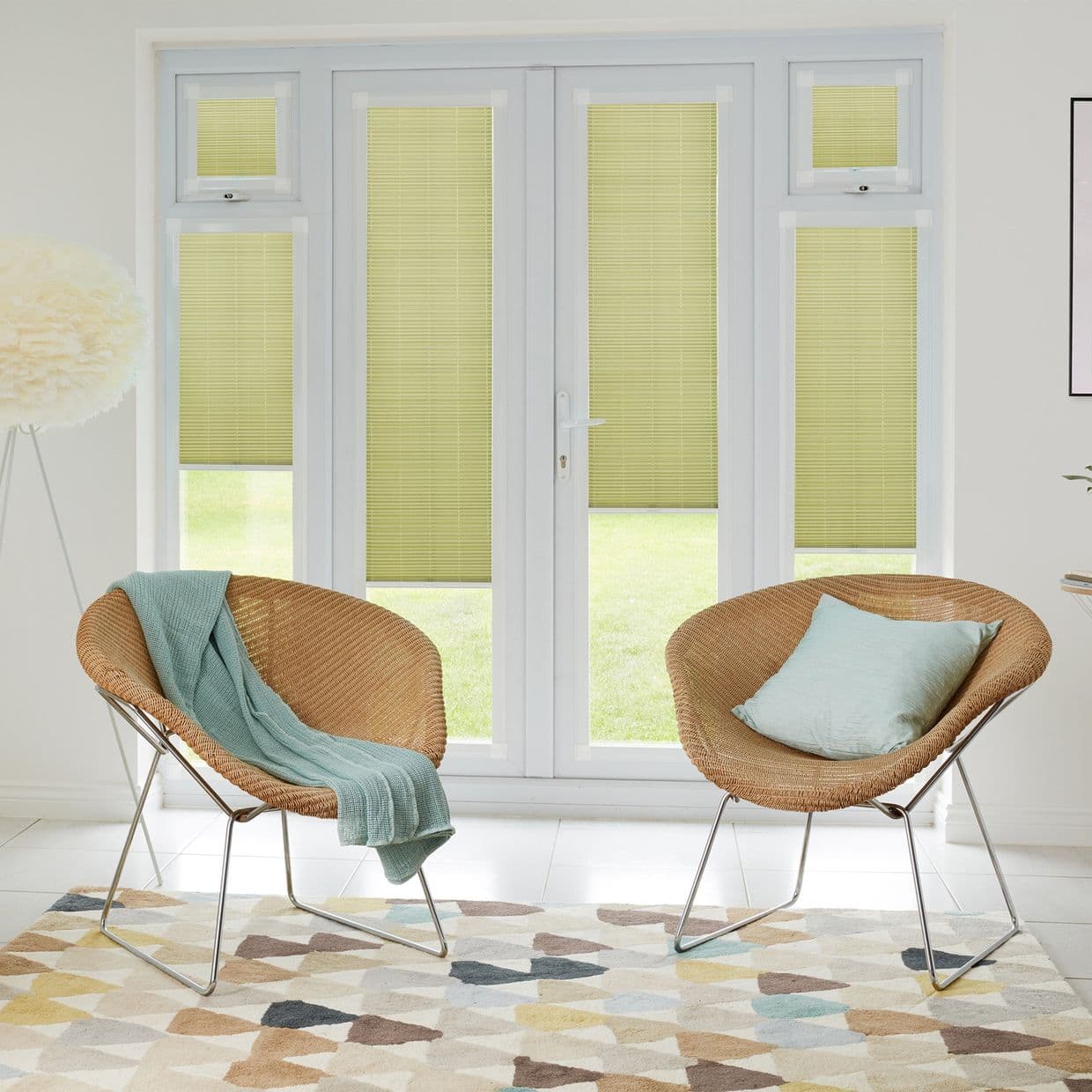 Yellow pleated blinds cover tall doors and sidelights, partially lowered and filtering soft daylight; in a bright living room with two wicker chairs, pale-blue textiles, a geometric rug, and a lawn view.