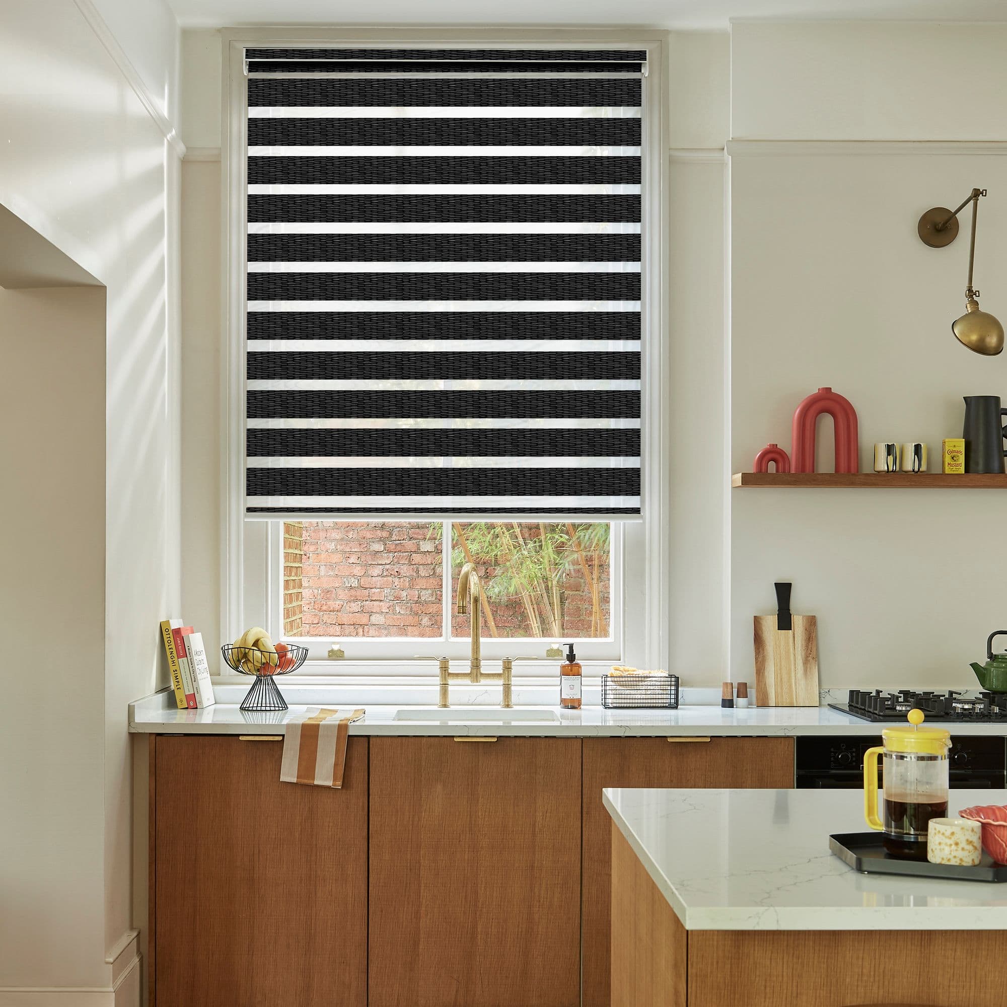 Black-and-white striped roller blind, lowered with alternating opaque and sheer bands, filtering light and partially obscuring a window above a kitchen sink with brass faucet, countertop, wooden cabinets and brick outside.