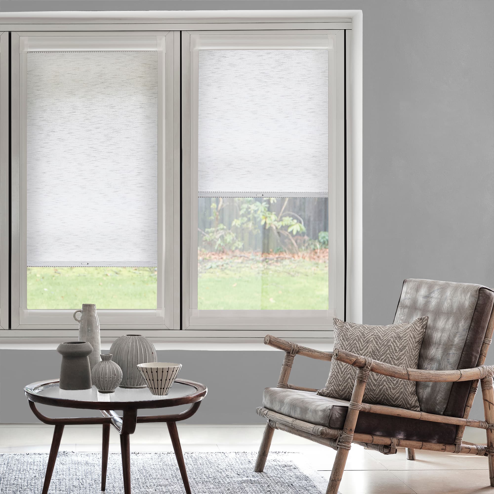 White textured roller blinds, left fully lowered and right partially lowered, filtering daylight over two tall windows; contemporary gray living room with bamboo-frame leather armchair, coffee table, and garden view.