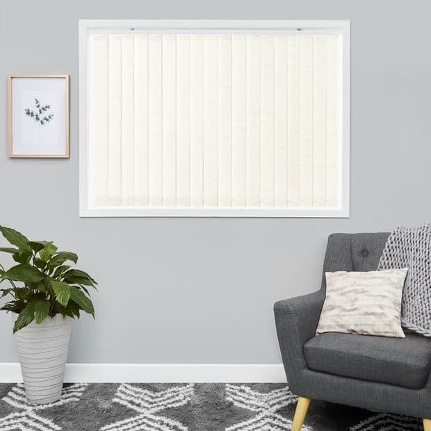 Cream vertical blinds fill a centered window, evenly spaced slats closed, filtering daylight; in a grey living room with a potted plant, framed botanical print, patterned rug and grey armchair.