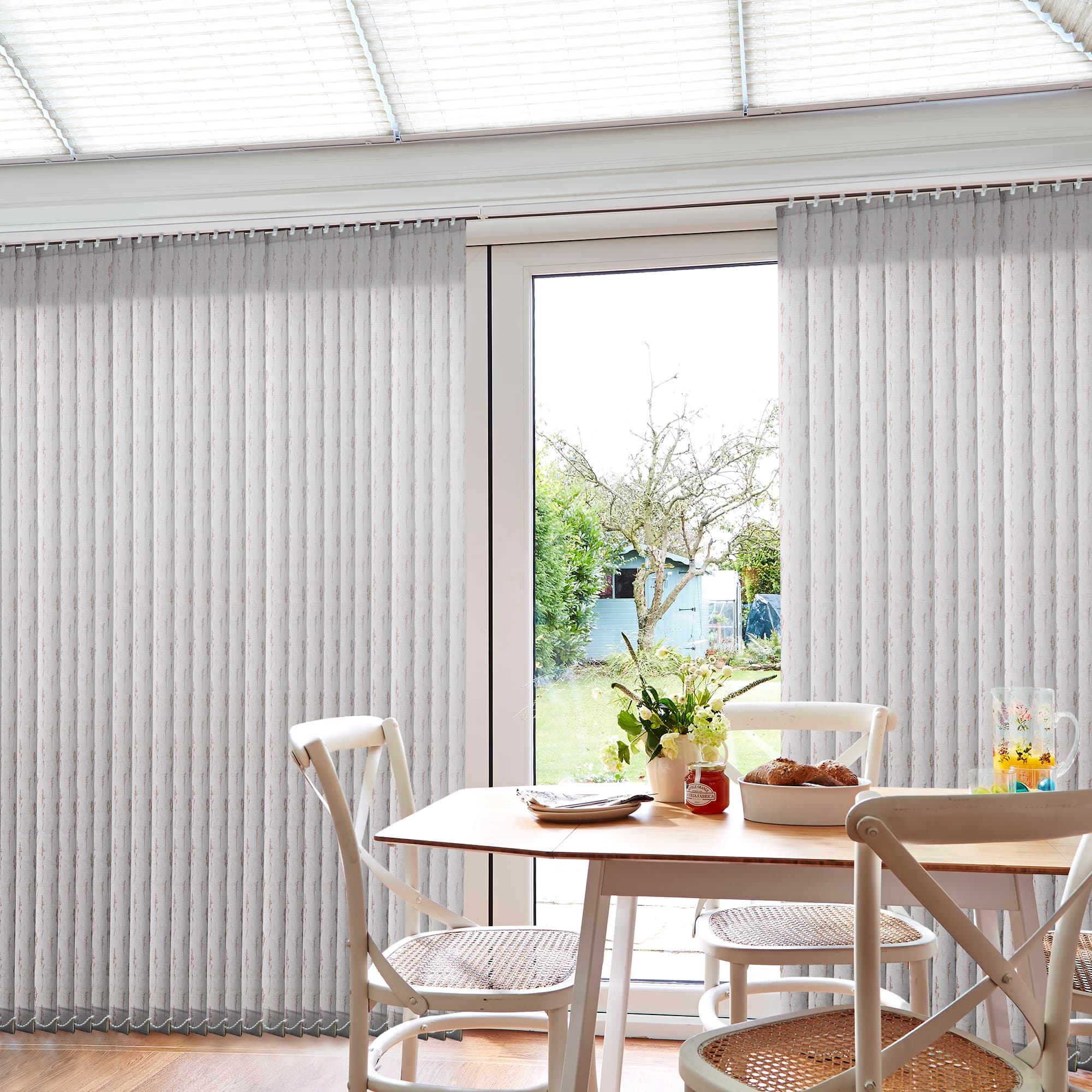 Light-gray textured vertical blinds cover sliding patio doors, slightly parted at center to reveal glass. They filter soft daylight over a small dining table, chairs and garden view.