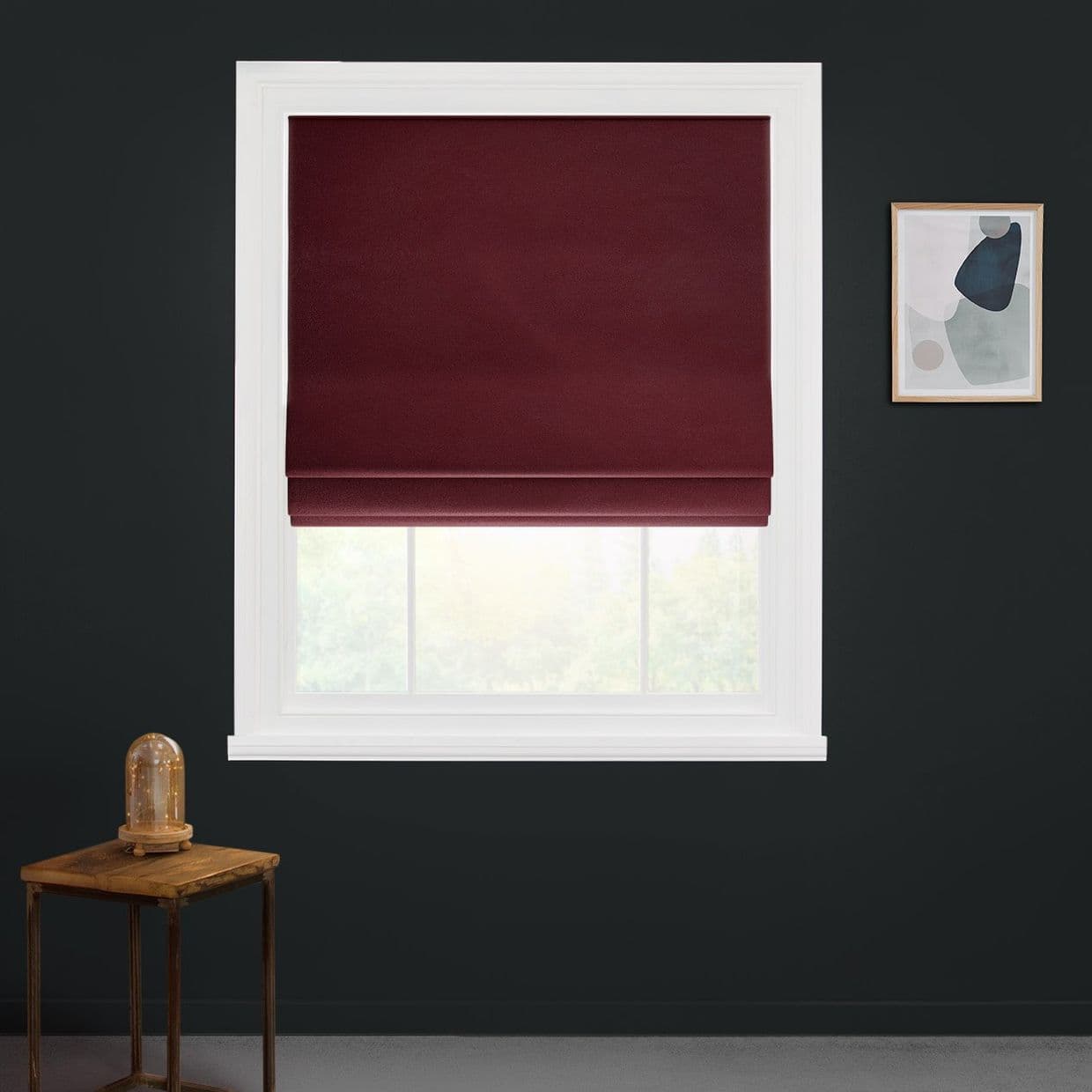 Burgundy Roman shade, lowered with folds, blocking most light; inside a white window frame, allowing faint daylight at the bottom, on a dark wall beside a small table and abstract art.