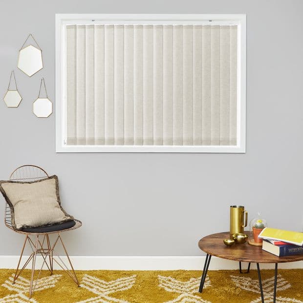 Beige fabric vertical blinds cover a recessed window, closed and filtering soft daylight; grey-walled living room with a rattan chair, round wooden table and mustard patterned rug.