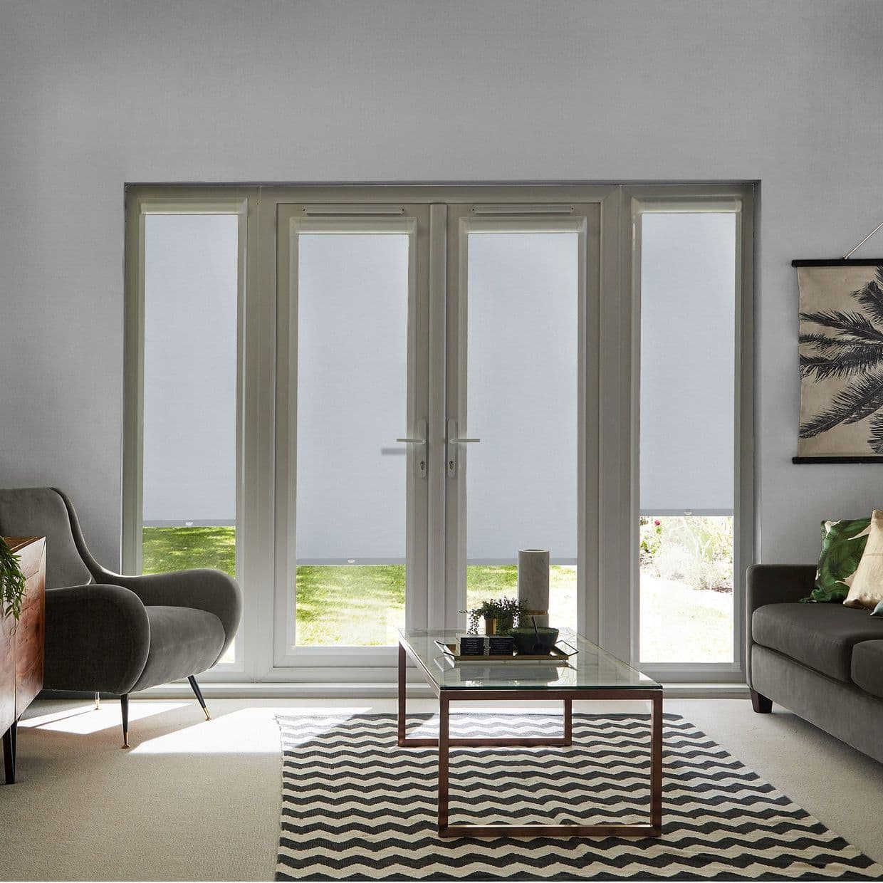 Four white roller blinds cover central French doors and sidelights, lowered almost fully with narrow bottom reveals, filtering daylight in a living room with glass table, chevron rug, armchair, sofa.