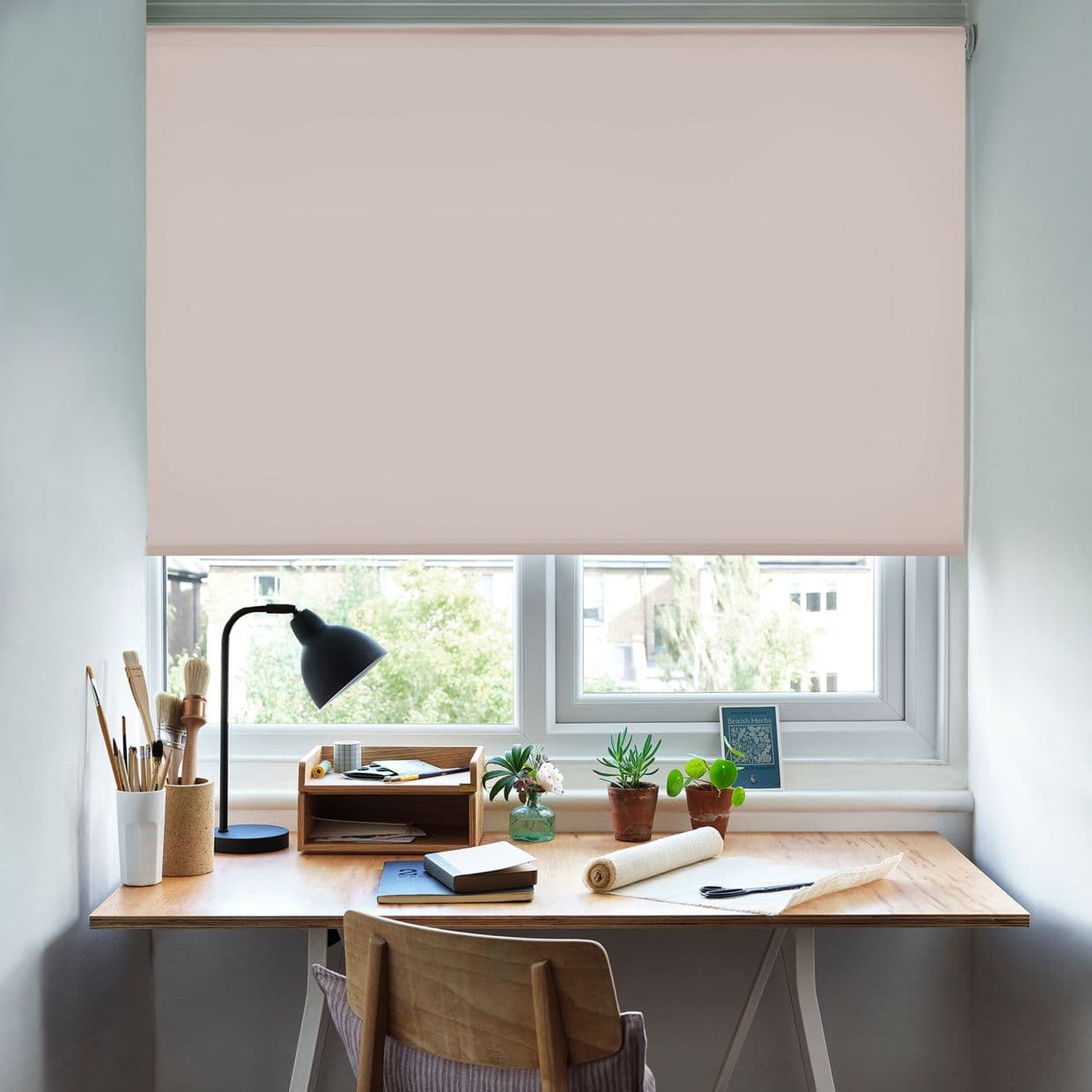 Pale blush roller blind, fully lowered across a doubleâ€‘pane window with smooth fabric, blocking and softly diffusing daylight; a small home desk beneath holds a lamp, plants, and brushes.