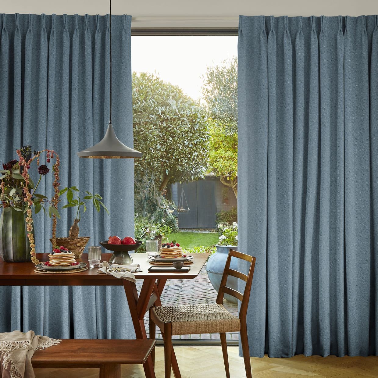 Full-length pleated blue curtains, parted slightly, letting soft daylight through a sliding glass door; a wooden dining table set with pancakes, a pendant lamp, and a lush garden view.