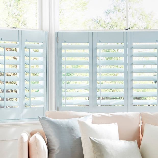 Pale blue plantation shutters across lower window panels, louvered and hinged, partially open, filtering soft sunlight into a bright living room with a cream sofa and pastel cushions, garden view.