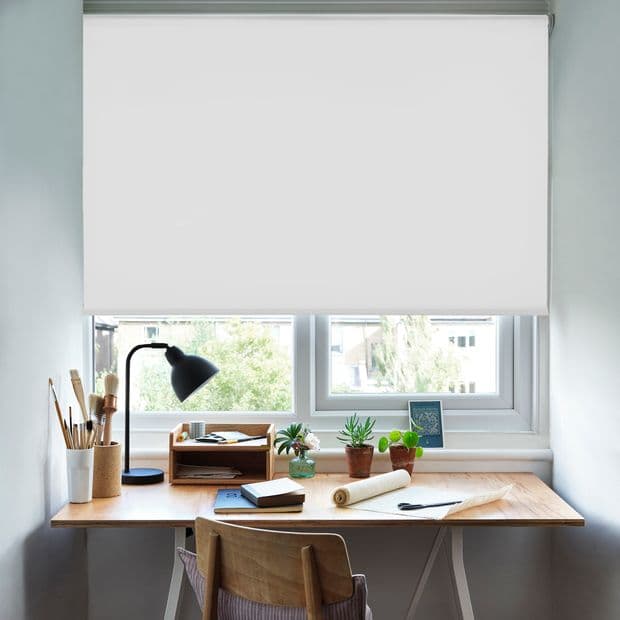 A white roller blind lowered over a double window, blocking light; beneath, a wooden desk with lamp, potted succulents and stationery in a bright, tidy home office with soft daylight.