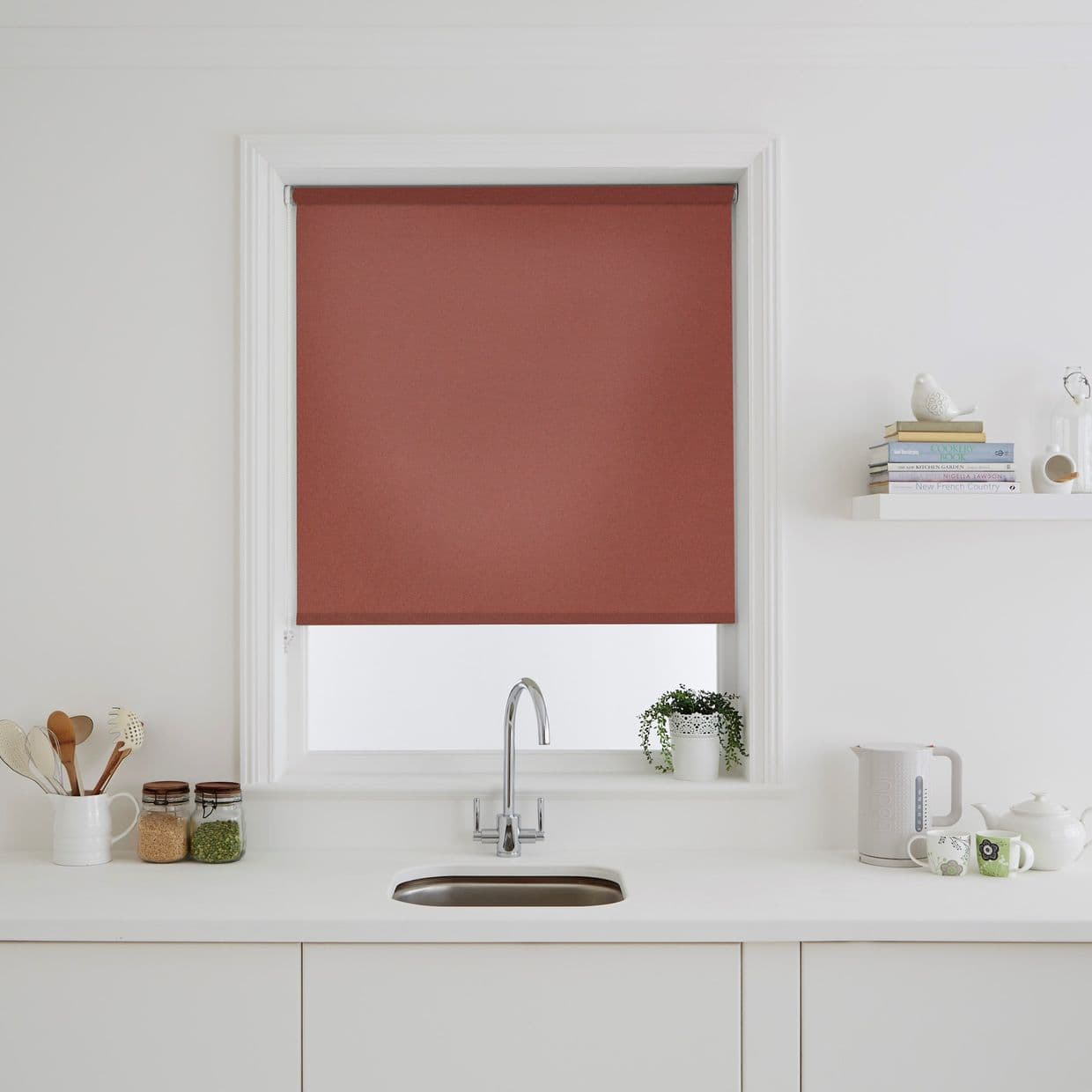 Terracotta fabric roller blind, lowered and covering the window, blocking view and light; above a white kitchen sink and countertop with jars, utensils, potted plant, kettle, and shelf.