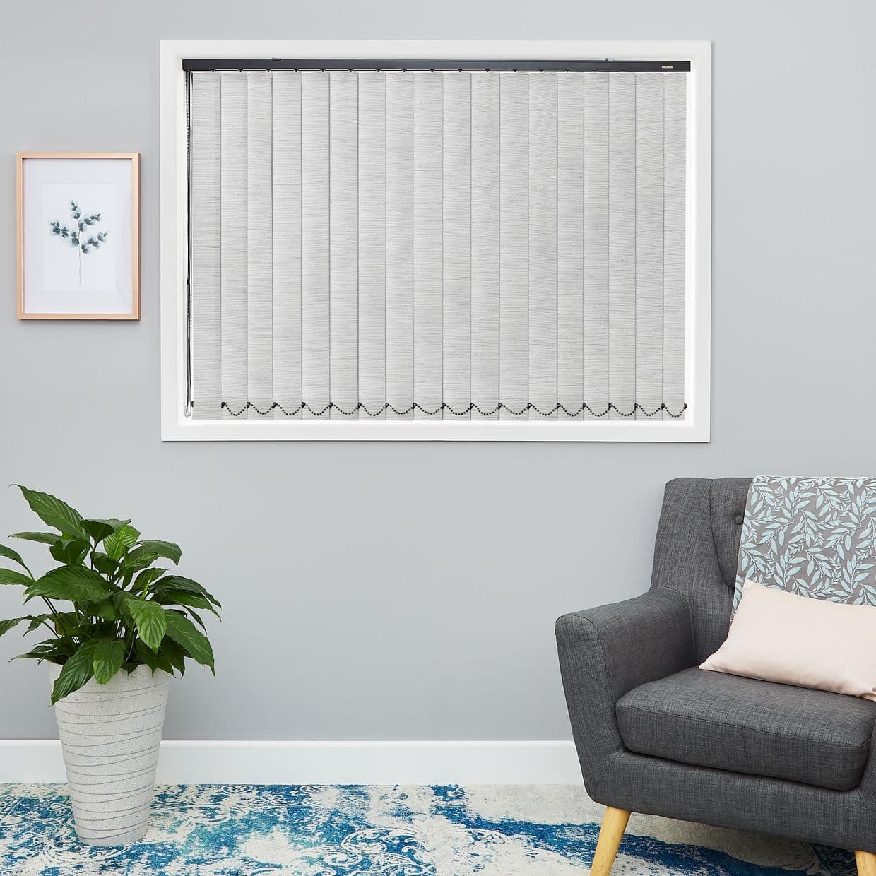 Vertical fabric blinds, fully closed across the framed window with scalloped bead chain along the bottom, filtering light; modern gray living room with armchair, potted plant and patterned rug.