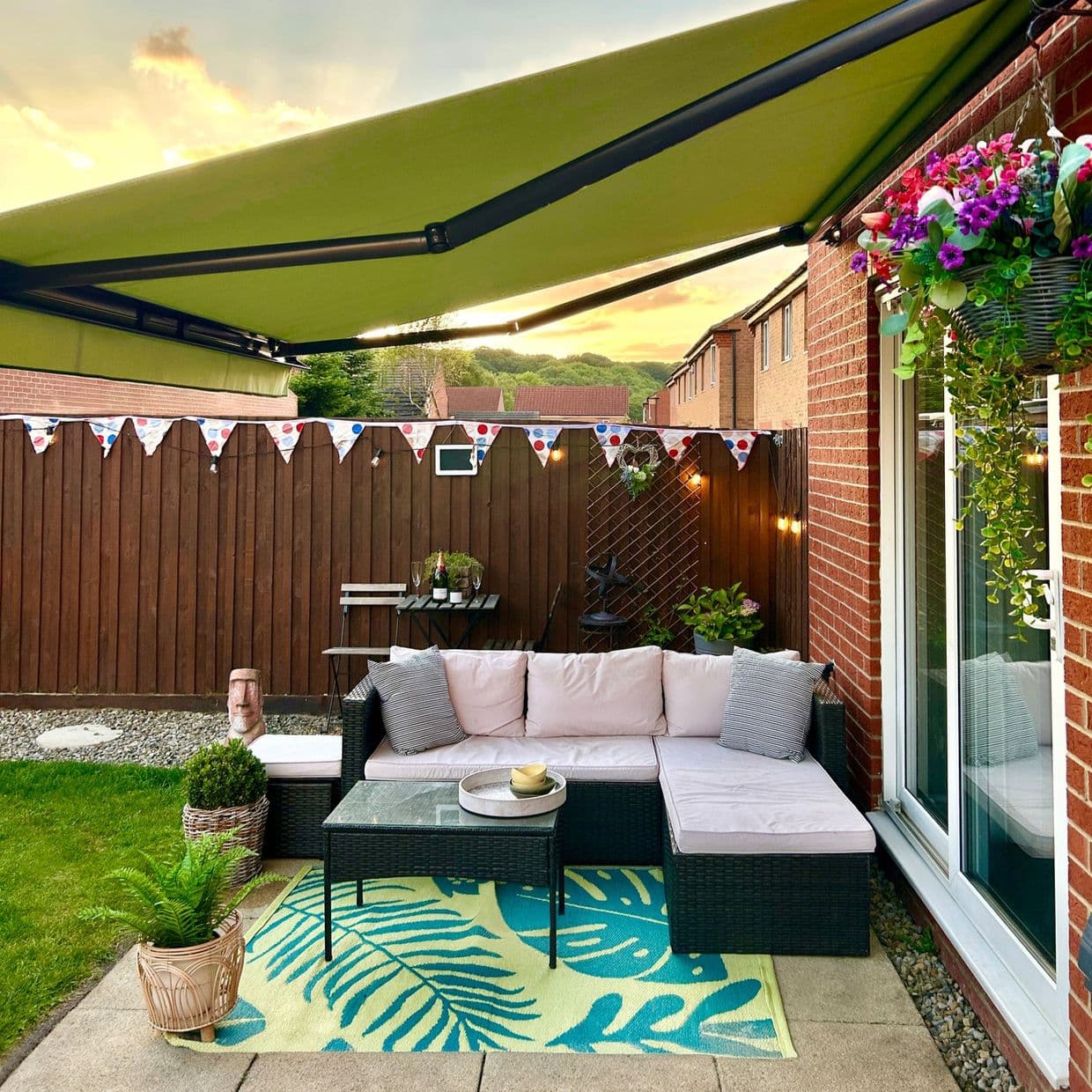 Green retractable awning with black metal arms, fully extended, shading a small backyard patio beside a brick house; wicker L-shaped sofa on tropical rug, hanging flowers, bunting at sunset.