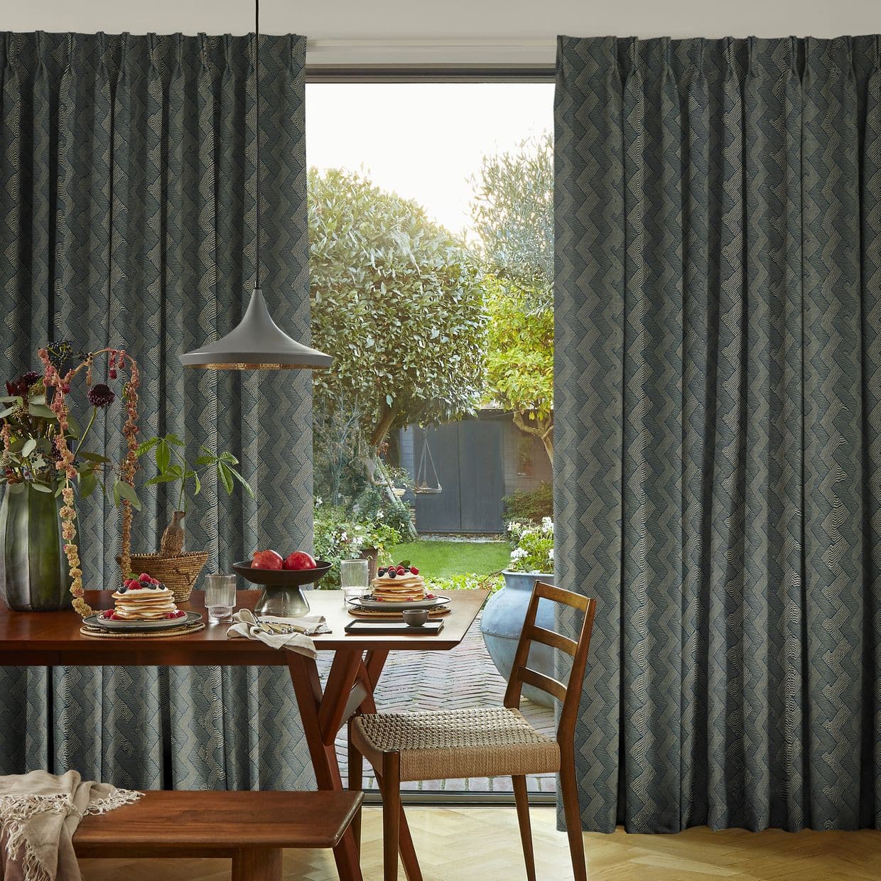 Floor-to-ceiling, patterned pleated curtains, dark blue chevron, partially drawn to frame a sliding glass door; filtering morning light over a dining table set with pancakes, pendant lamp, and backyard garden.