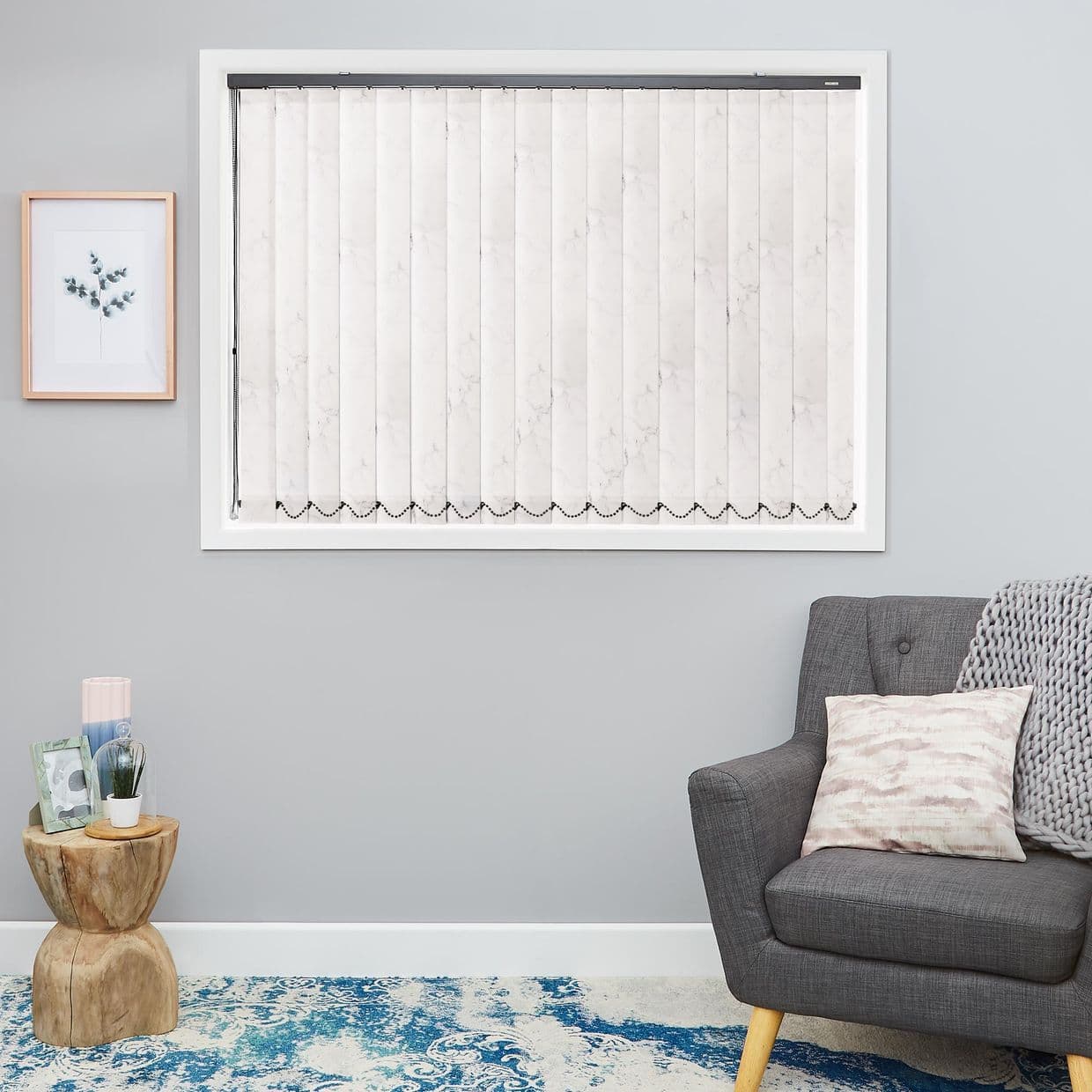 White marble-pattern vertical blinds in a white-framed window, closed and diffusing light; on a light-gray living-room wall beside a framed botanical print, stump side table, gray armchair and blue rug.