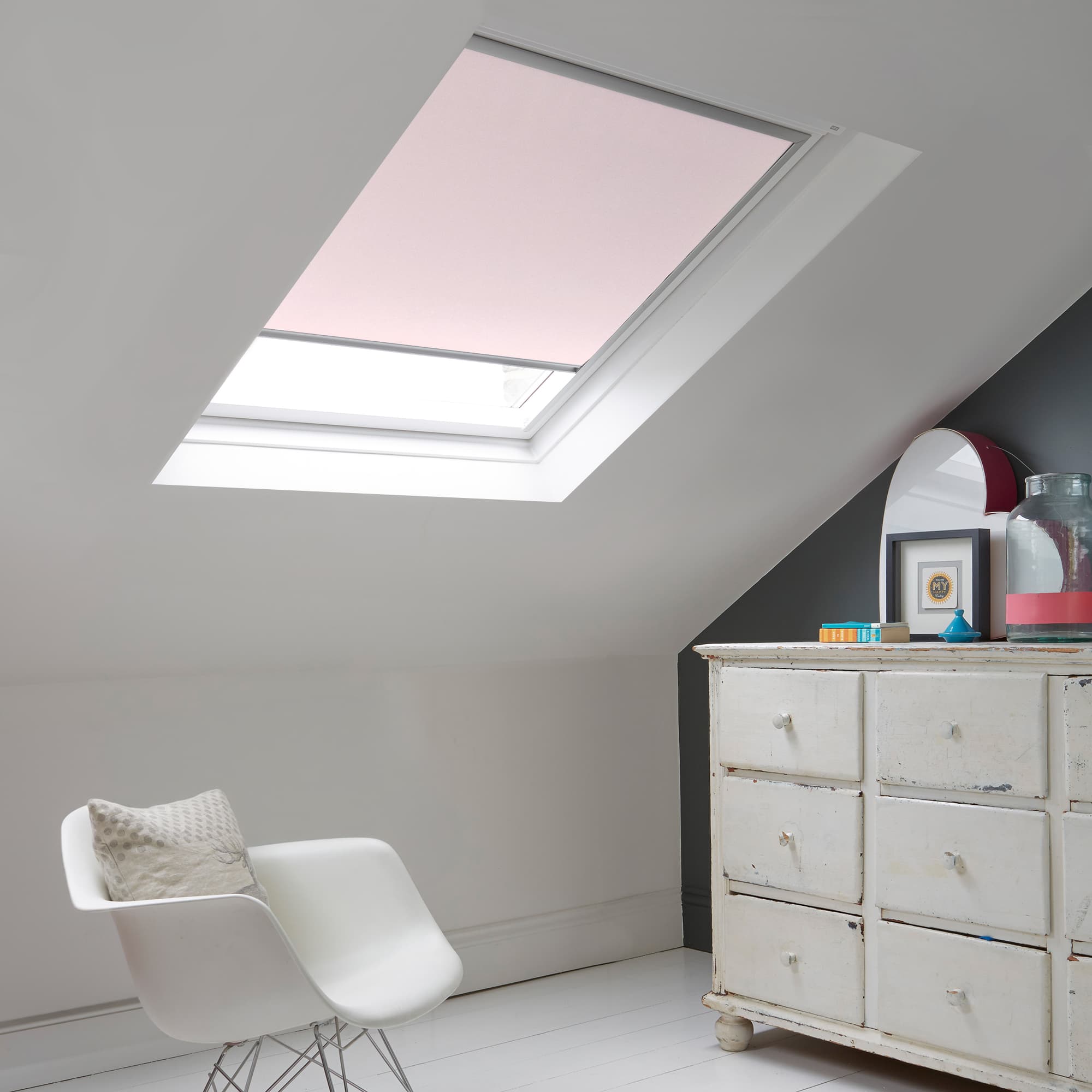Room with white eaves wall, dark blue-grey wall to right with a shabby chic white drawer unit, a contemporary white chair, white painted floorboards and a skylight blind in pale coral.