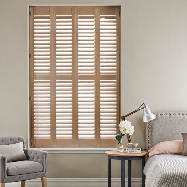 Full-height wooden louvered shutters covering a recessed window, narrow panels with slats tilted to admit diffused light; in a softly lit bedroom beside an upholstered bed, table and chair.
