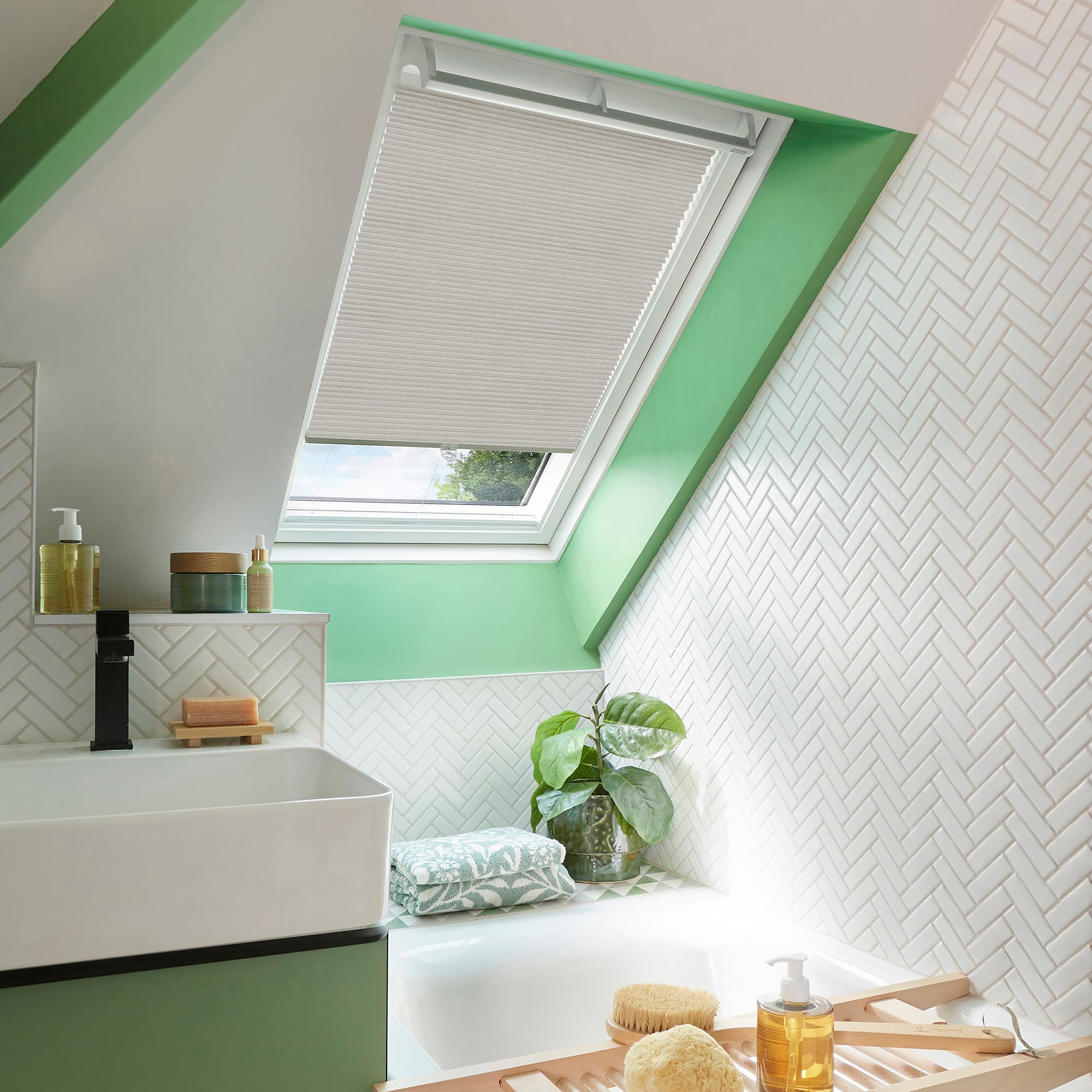 Beige pleated skylight blind, lowered over most of the roof window leaving a small bottom gap, filtering light into a bright bathroom with mint trim, white herringbone tiles and bathtub.