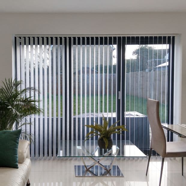 Floor-to-ceiling vertical blinds with narrow slats cover sliding glass doors; mostly closed, they filter daylight and reveal a fenced grassy yard beyond a bright tiled living area.