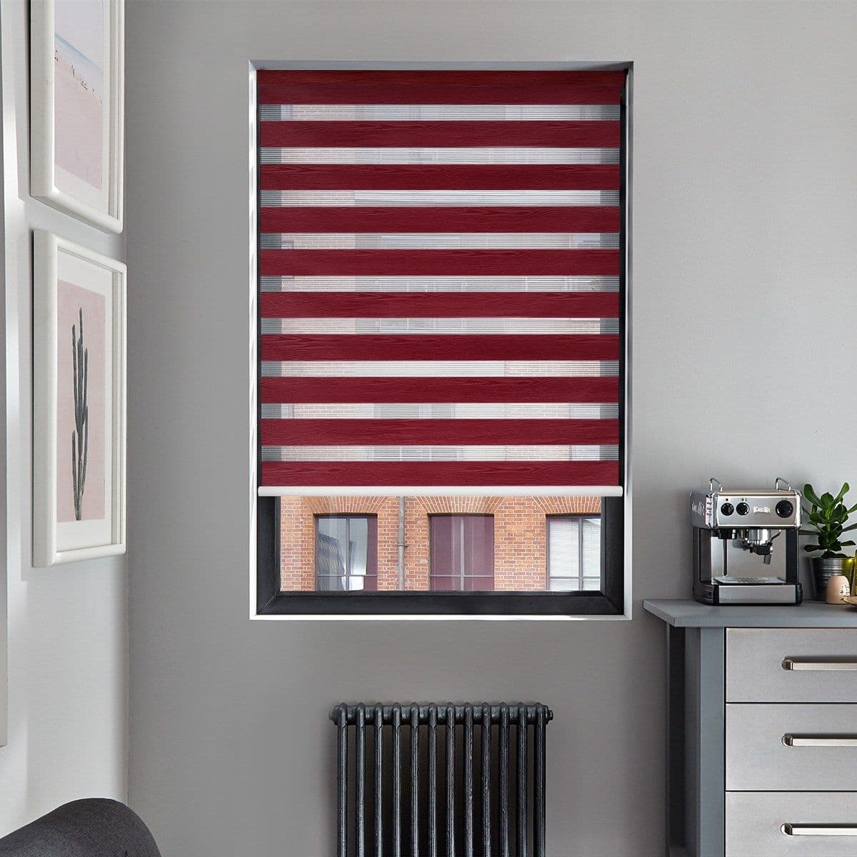 A burgundy-and-sheer striped zebra roller blind, lowered across the window with alternating horizontal bands, filters light and partly obscures a brick-building view in a gray kitchen corner with espresso machine.