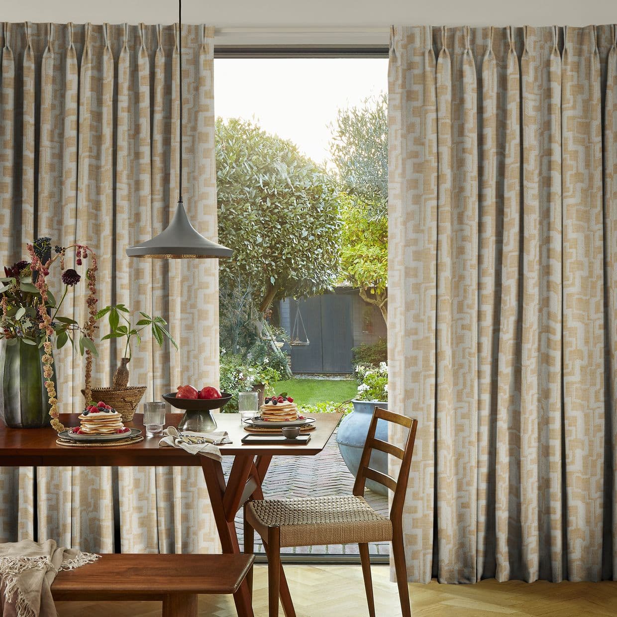 Pleated beige geometric curtains, hung full-length and parted at the center, frame a glass door and filter daylight; a dining table with plates and pendant lamp inside, garden visible outside.