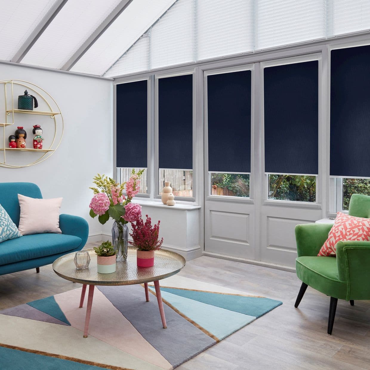 Dark navy roller blinds, lowered across tall conservatory windows, covering upper panes and blocking light; a living room contains a teal sofa, green armchair, pastel rug, coffee table with flowers.