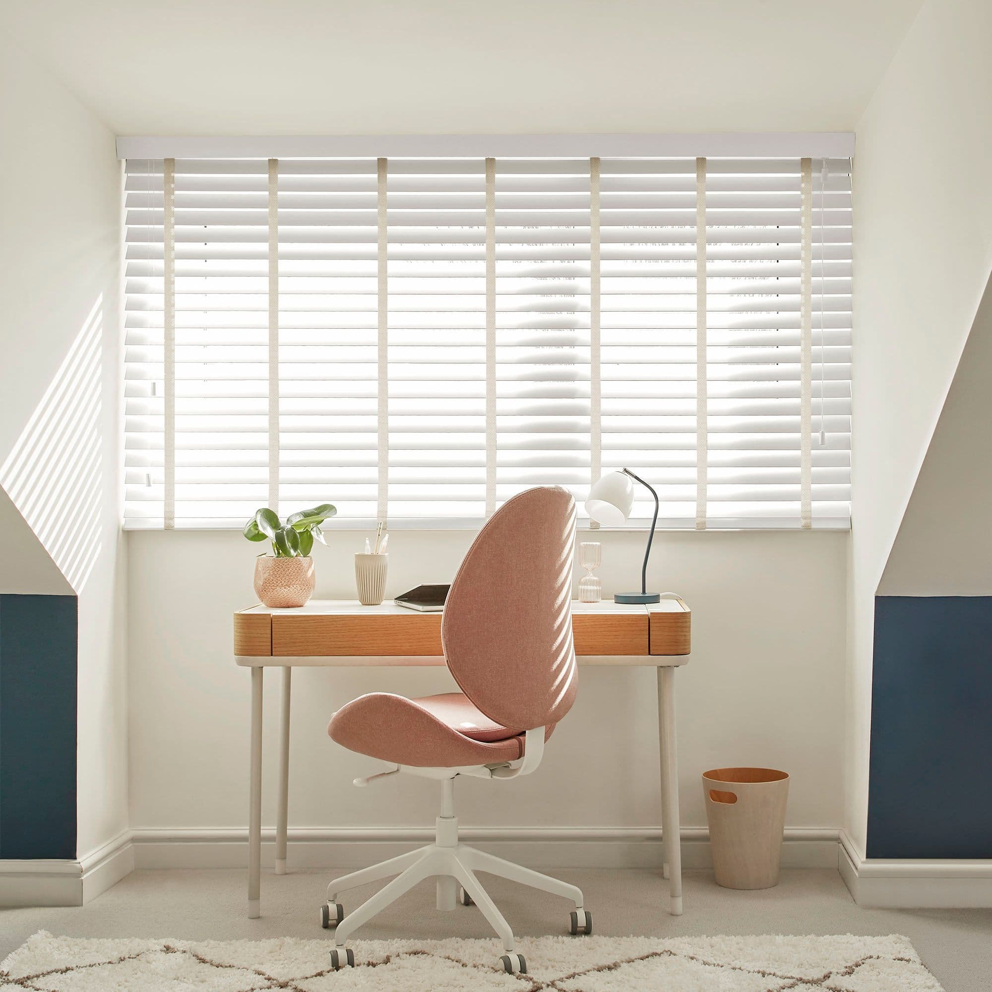 White horizontal slatted blinds with beige ladder tapes covering a dormer window â€” closed, filtering bright daylight â€” above a compact home office: wooden desk, pink swivel chair, lamp, plant, neutral walls.