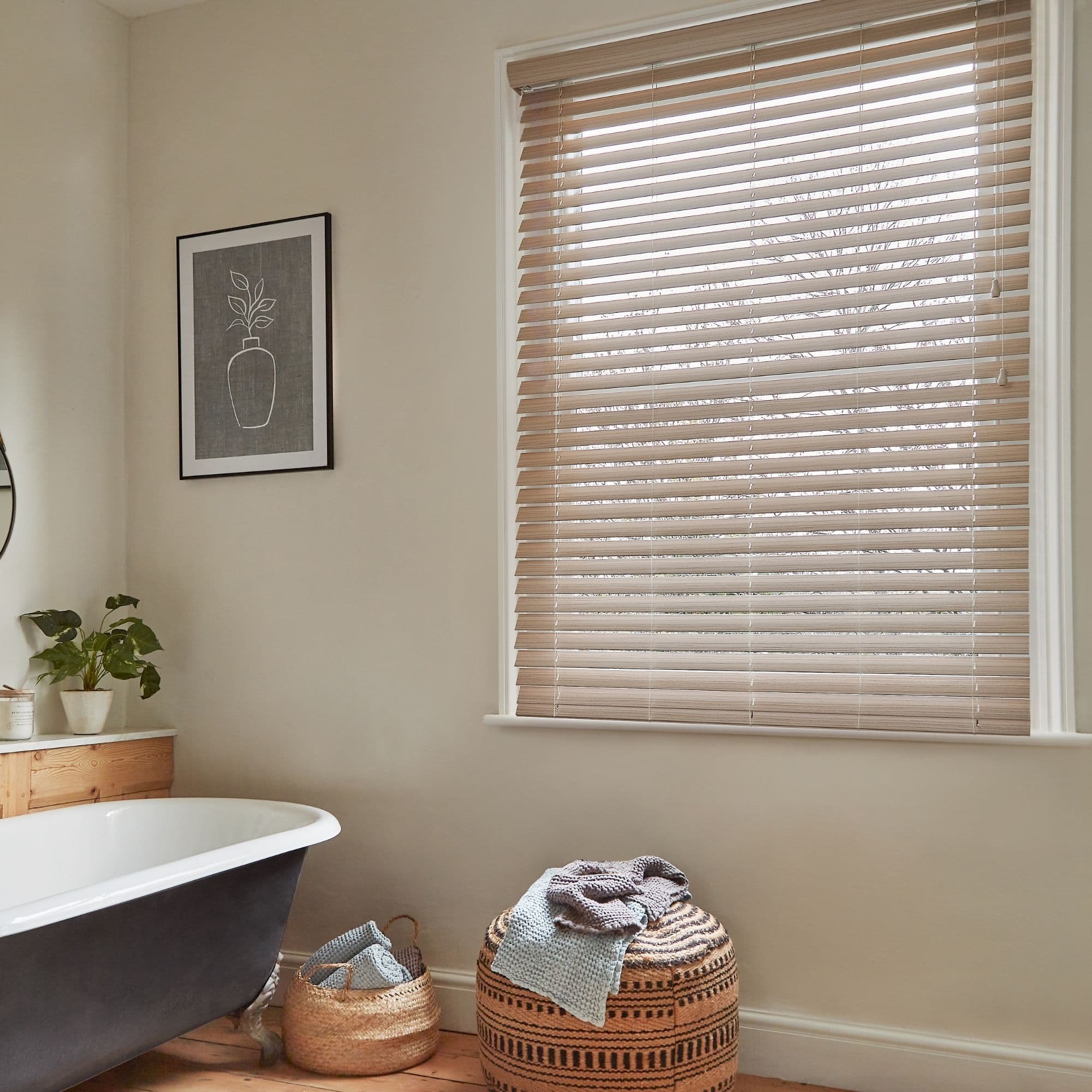 Wooden horizontal blinds covering a large window, slats tilted closed and filtering soft daylight; set above a neutral bathroom with clawfoot bathtub, potted plant, woven baskets, and patterned pouffe.