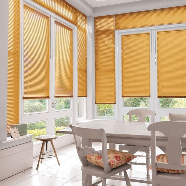 Honeyâ€‘orange horizontal slatted blinds cover tall bay windows and French doors, mostly lowered and filtering sunlight; bright dining area with white table, chairs with floral cushions and garden view.