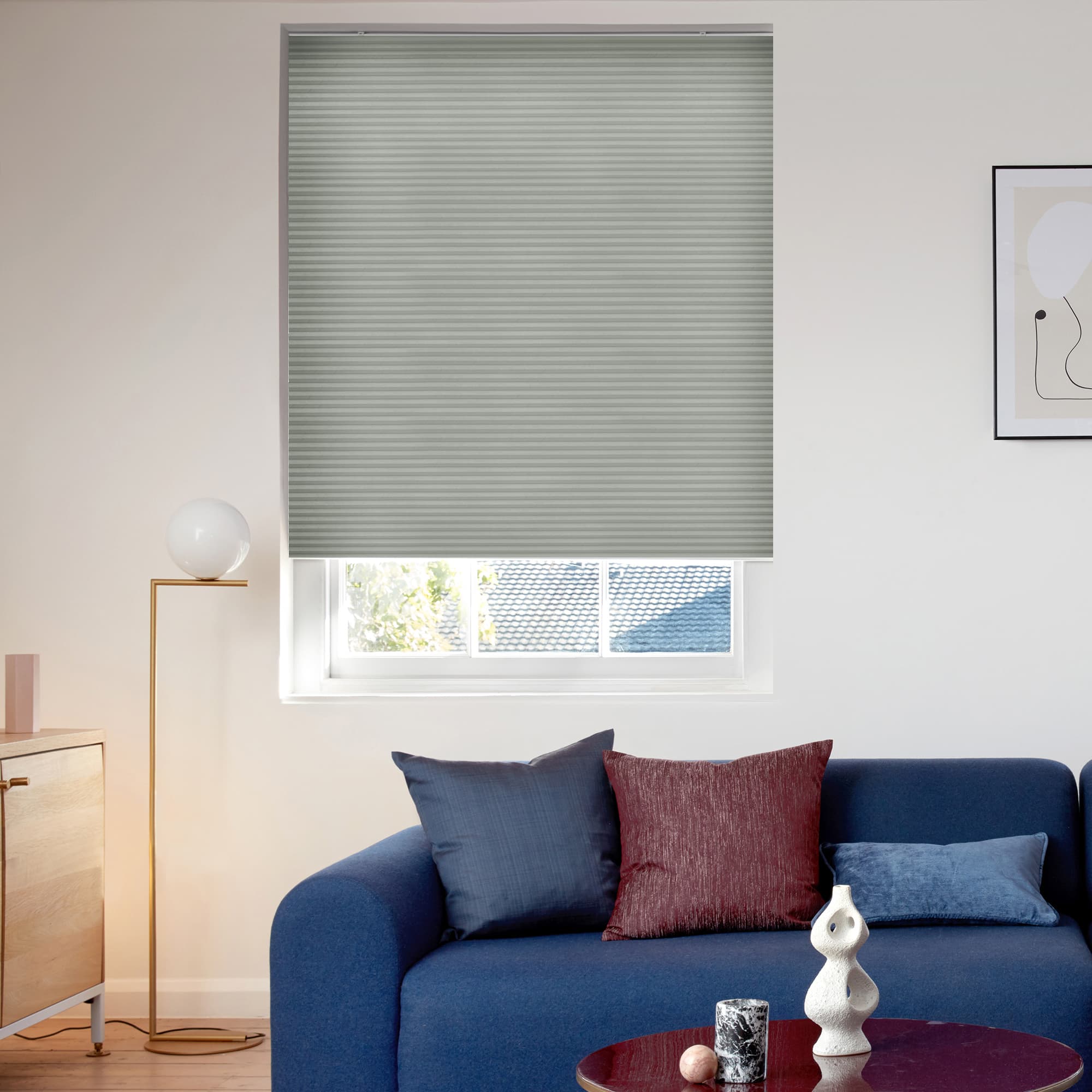 Pleated grey cellular blind, lowered to just above the sill with horizontal ribs, blocking most light; living room with blue sofa, cushions, floor lamp, framed art, roof and trees outside.