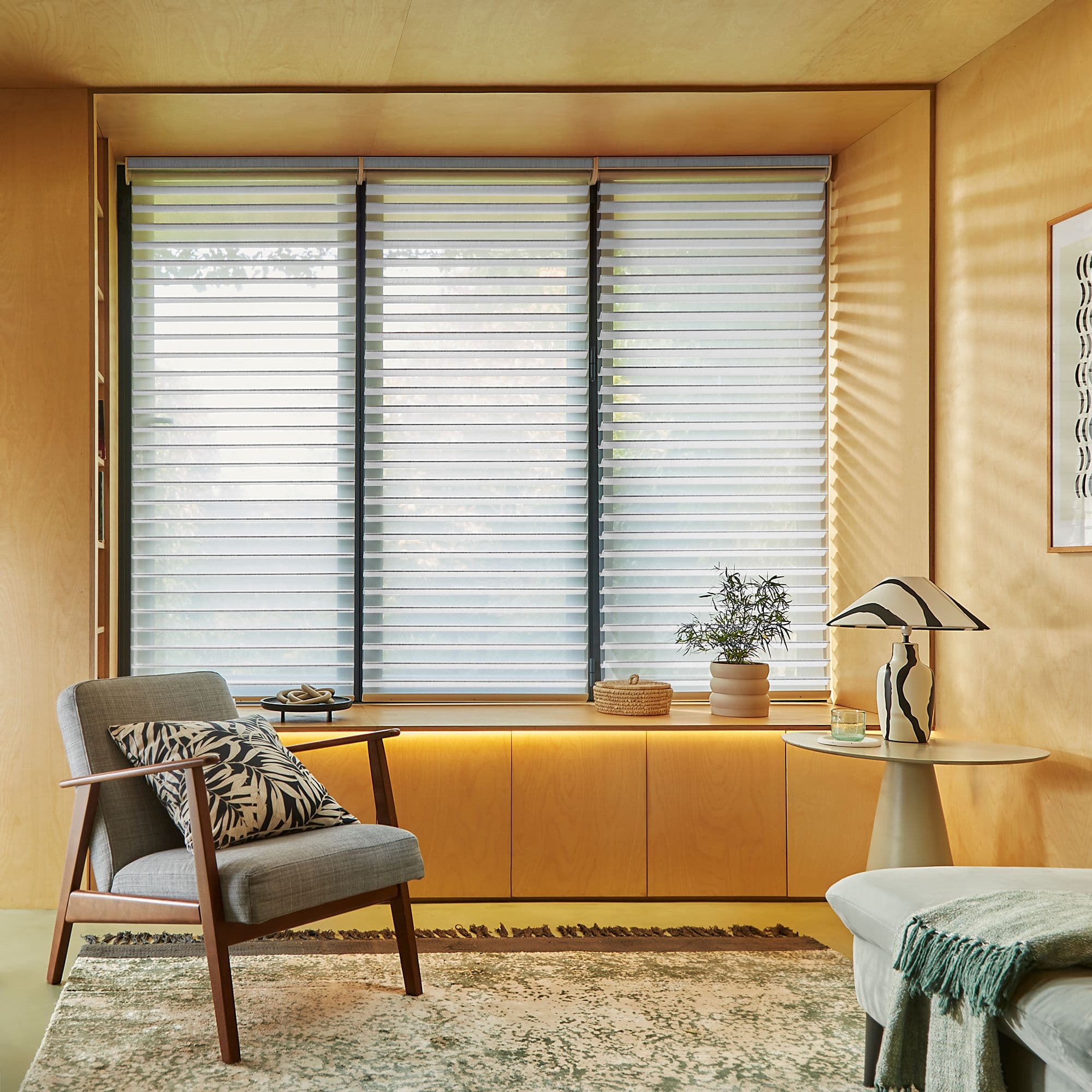 Layered horizontal blinds covering three tall windows, with alternating sheer and opaque bands; partially closed, filtering soft daylight into a warm wood-paneled living room with armchair, side table, and plant.