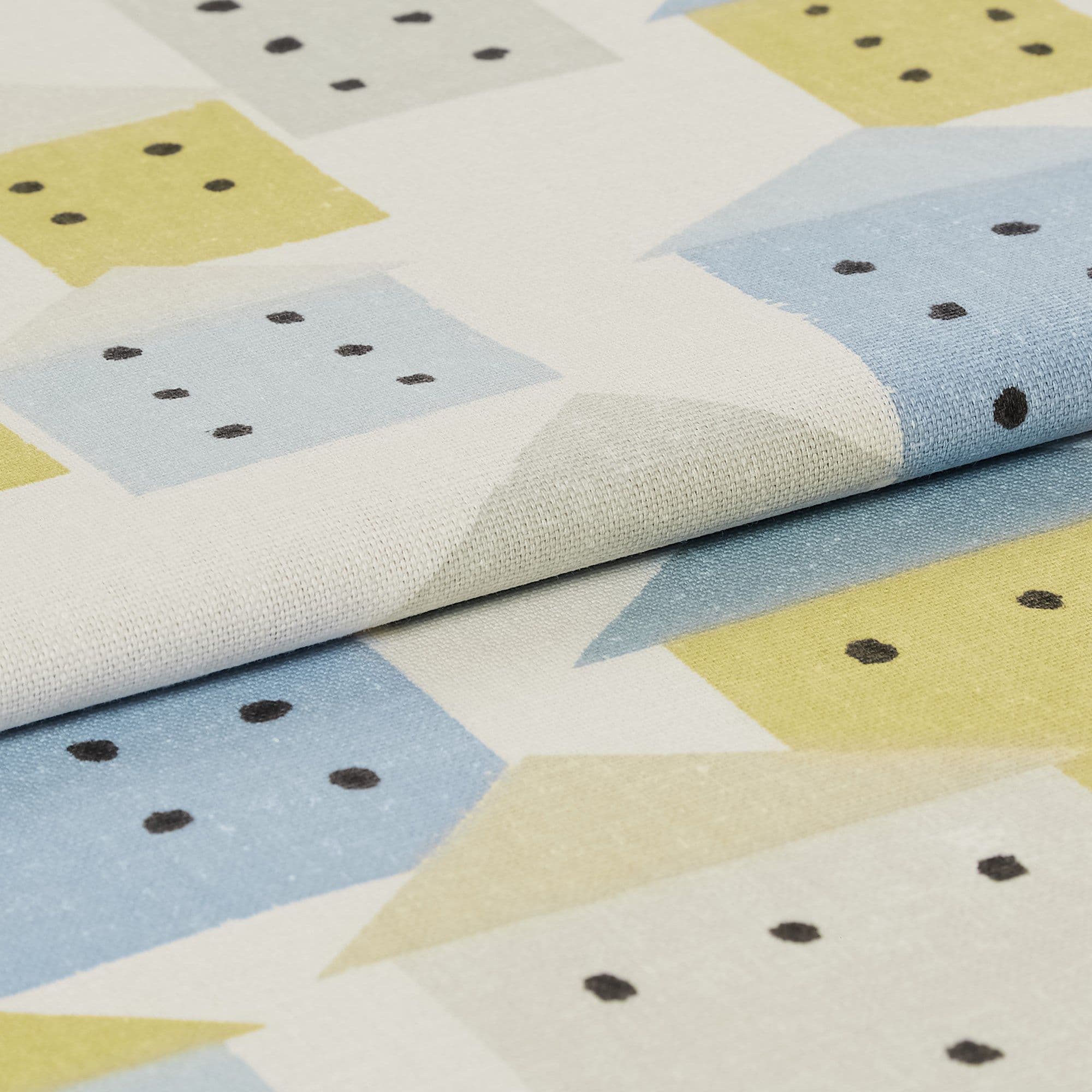 Folded printed cotton fabric showing pastel house-like blocks (blue, yellow, beige) with black dot "windows"; fabric lies layered, studio-lit close-up revealing woven texture.
