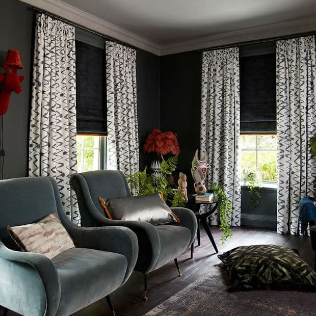 White patterned floor-length curtains flank three windows, paired with partially lowered black Roman shades that filter daylight; a dark-painted living room with velvet armchairs, plants and eclectic decorative objects.