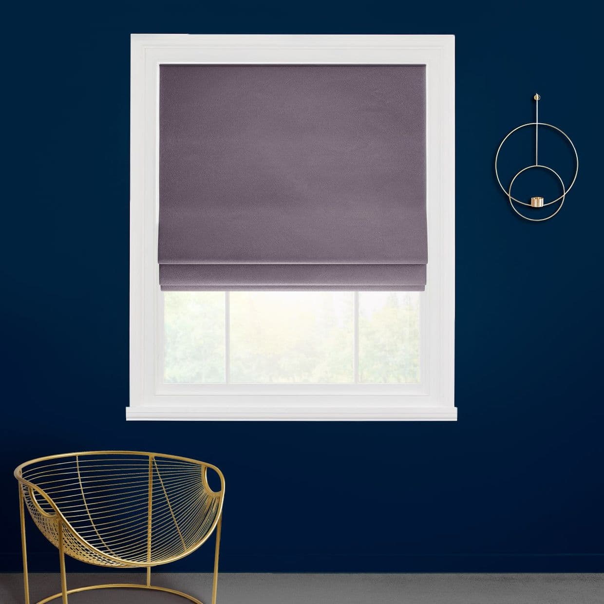 Mauve-gray fabric Roman shade lowered over a white-framed window, smoothing folds and filtering light; set against a deep navy wall with a gold circular sconce and gold wire chair.