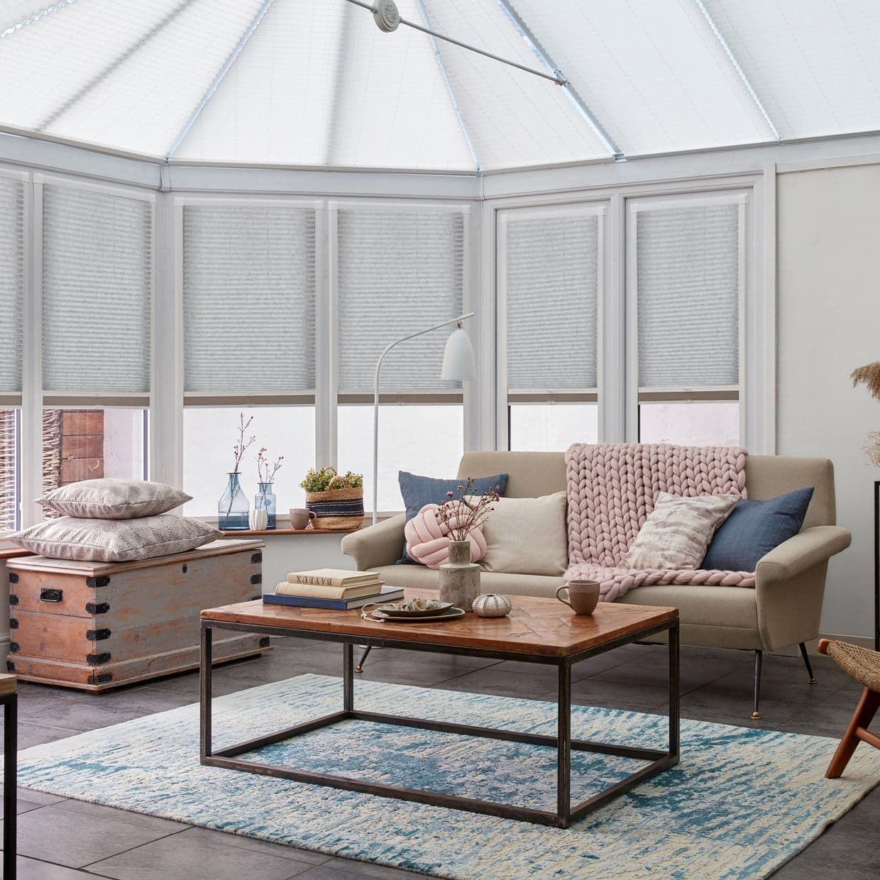 Light-gray pleated cellular blinds covering conservatory windows and roof, lowered and closed, softly diffusing daylight over a cozy sunroom with a sofa, wooden coffee table, and patterned rug.
