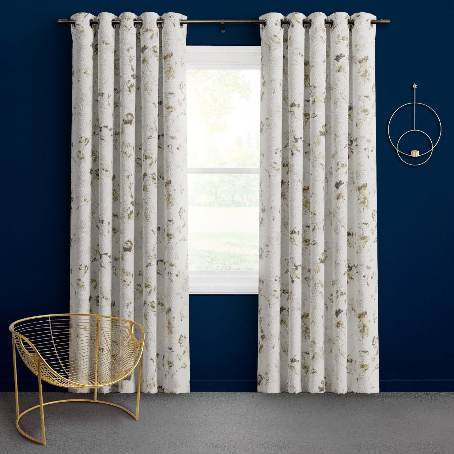 Grommet-top white floral curtains, pleated and hanging on a metal rod, frame a window and filter daylight; against a deep navy wall with gold wire chair on gray carpet.