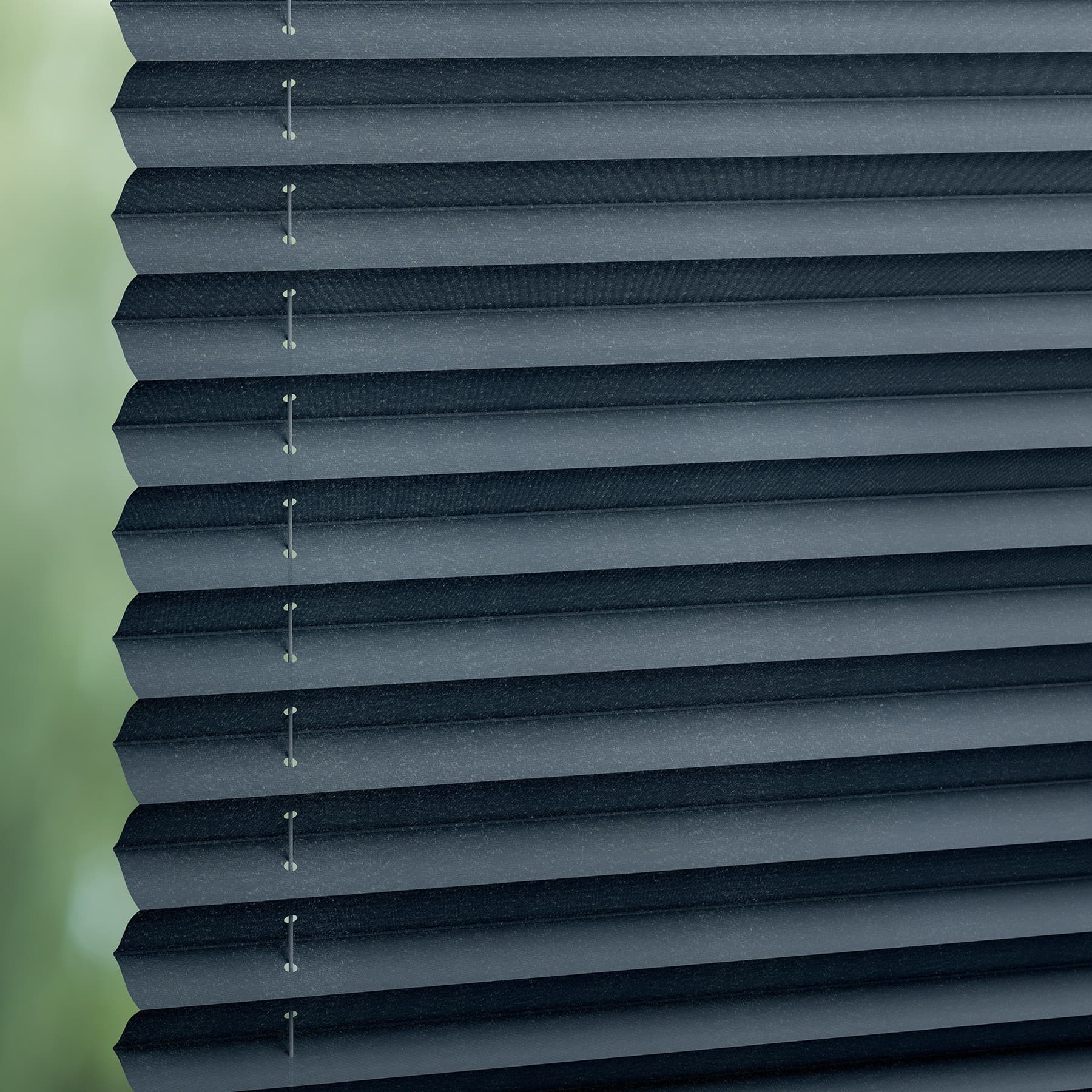 Dark gray pleated cellular blinds, stacked closed with visible cords and staples, blocking light; filtering soft daylight from a blurred green outdoor view through a window, creating diffuse interior lighting.