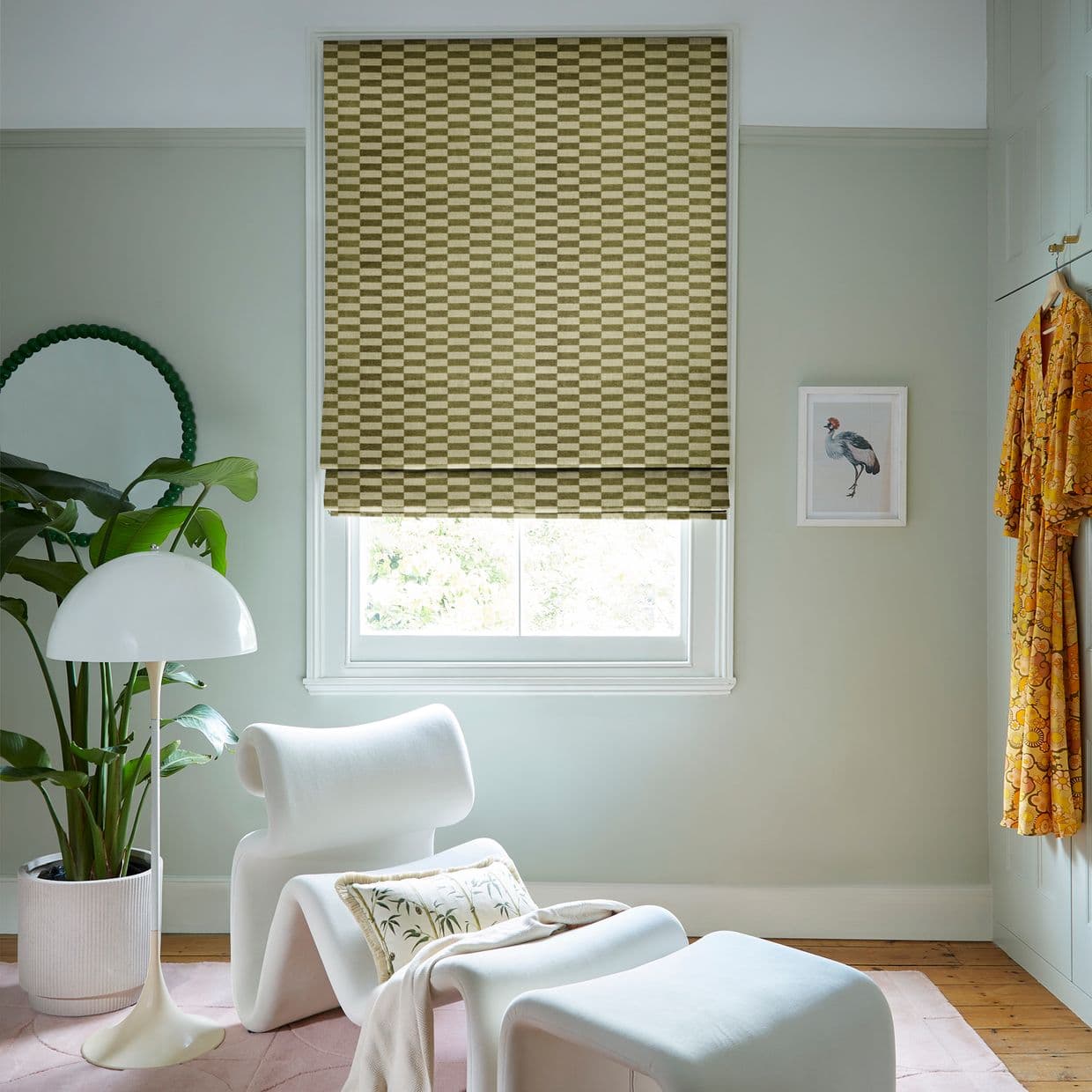 Checkered Roman blind covering the window, partially lowered and filtering daylight; in a pale-green sitting nook with a white lounge chair and ottoman, floor lamp, potted plant, framed bird print, hanging yellow robe.