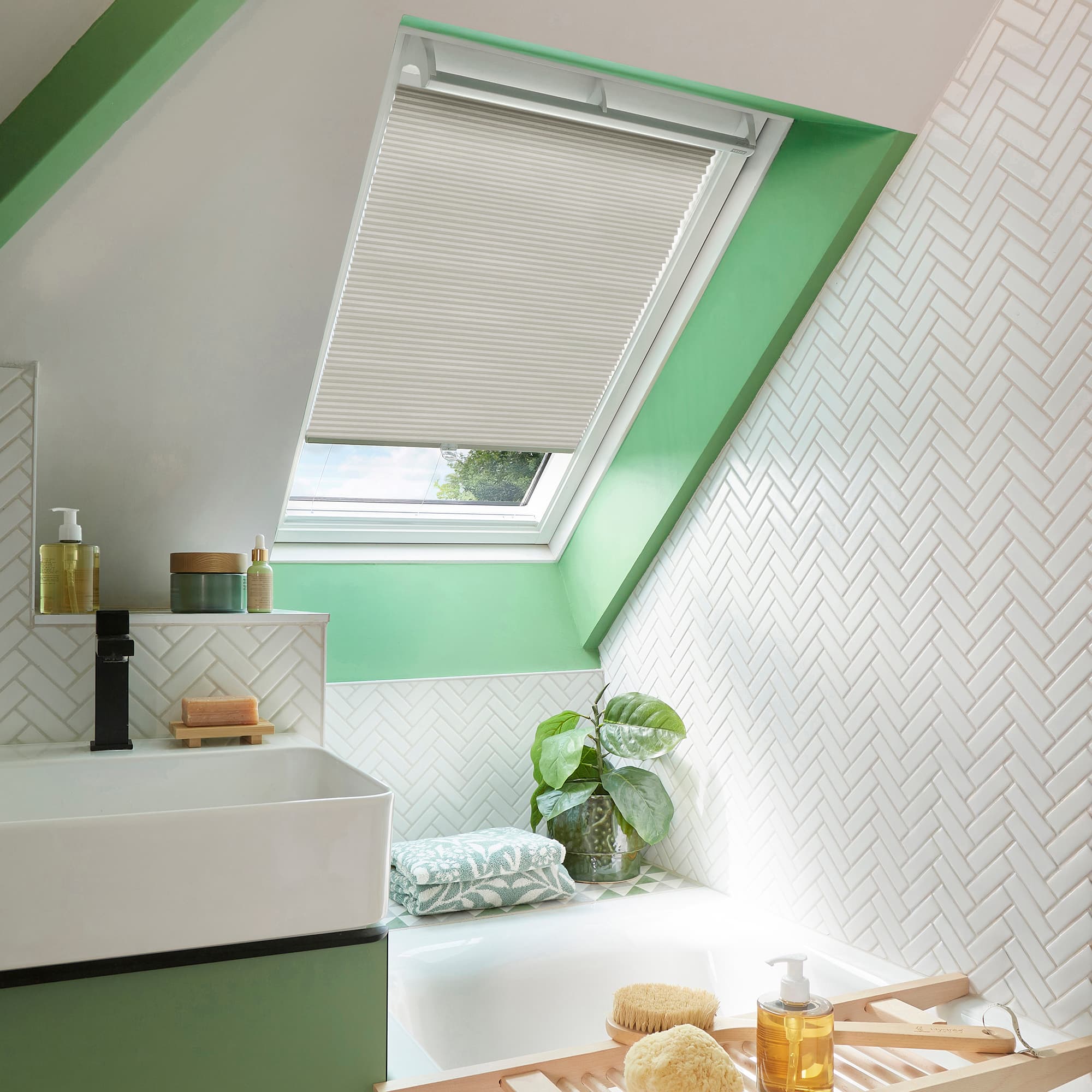 White pleated skylight blind, lowered halfway across a tilted window, filtering soft daylight; set in a greenâ€‘trimmed attic bathroom with white herringbone tiles, sink, tub, and a potted plant.
