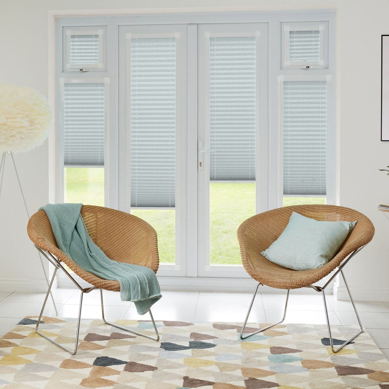 Light-gray pleated blinds cover French doors and sidelights, partially lowered and diffusing daylight; bright living room with two rattan chairs, mint throw and cushion, geometric rug, and lawn view.